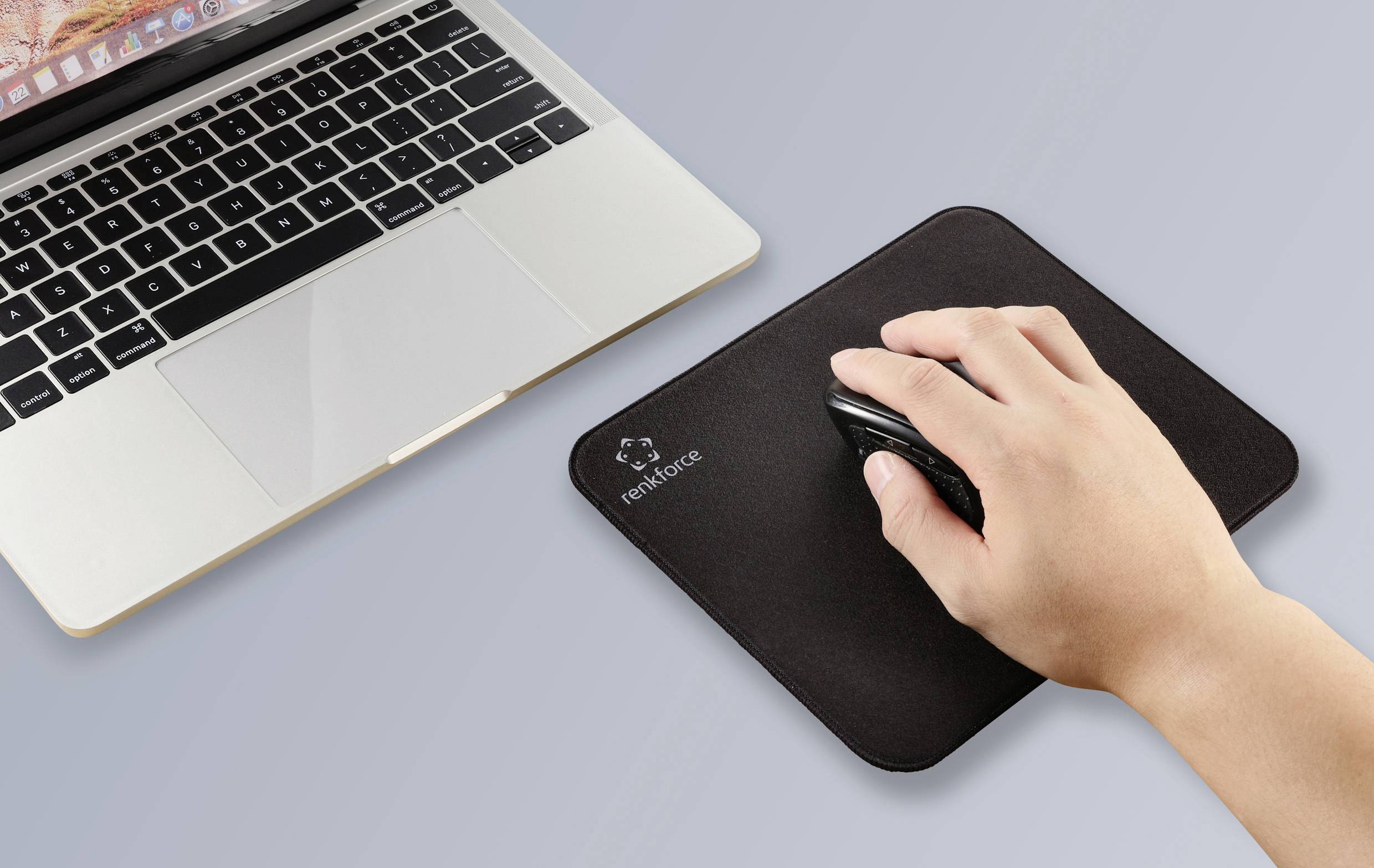 Ruka obsluhuje bezdrôtovú myš na čiernej podložke pod myšou vedľa laptopa na sivom povrchu.