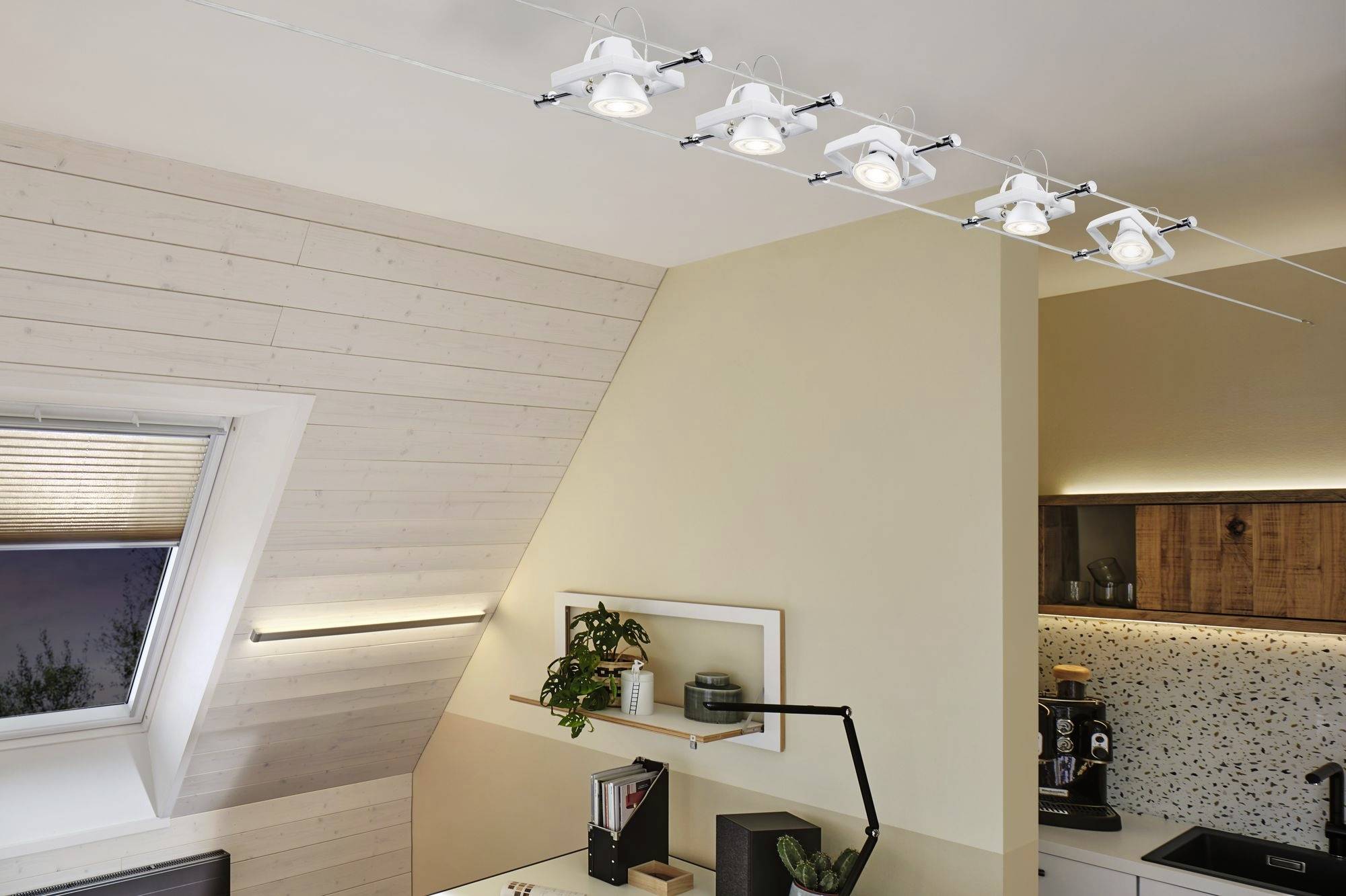 Moderná kuchyňa so šikmým dreveným stropom, osvetlená radom stropných svetiel. Polica s rastlinami a knihami.