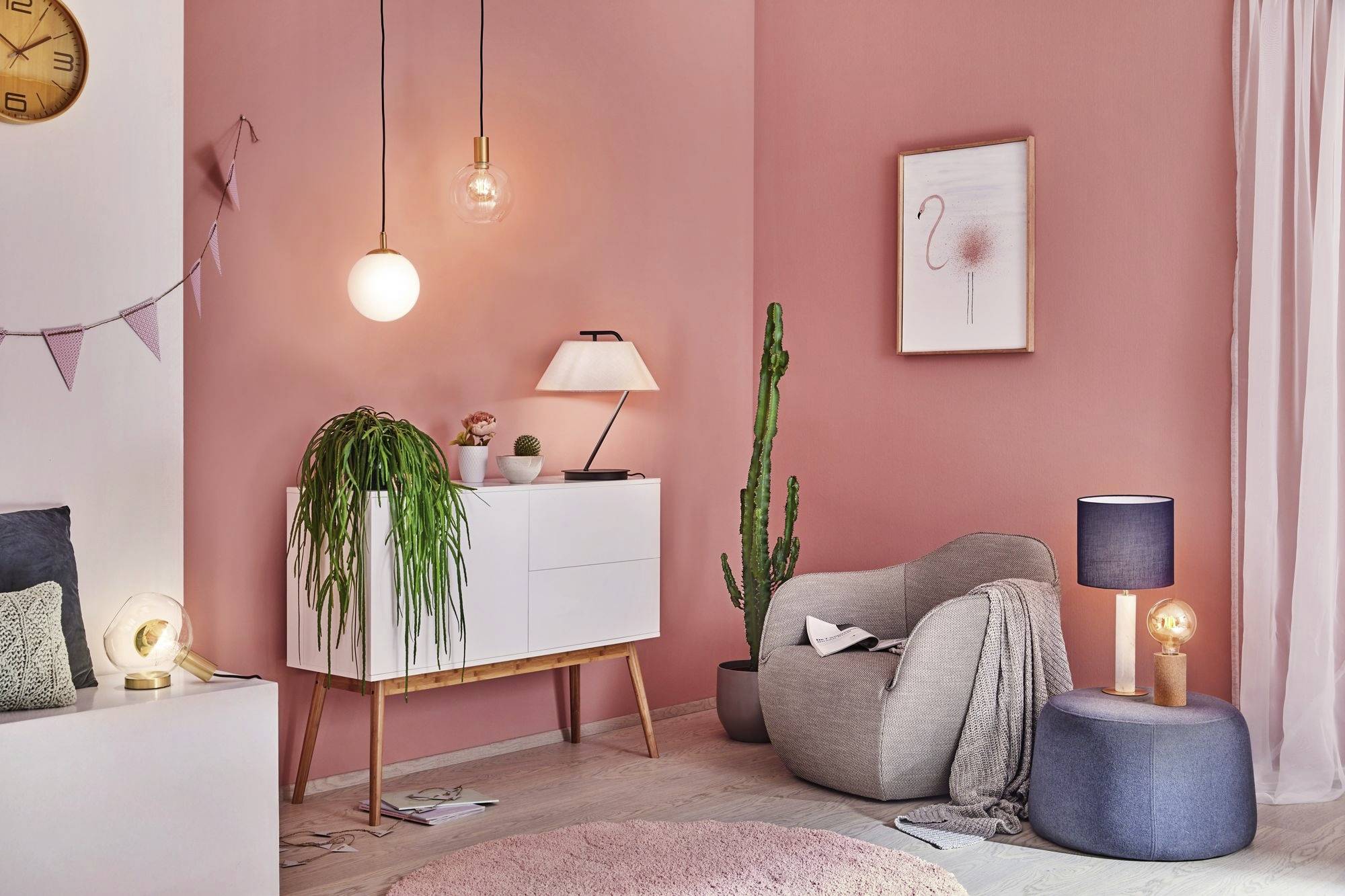 Útulná obývačka s ružovými stenami. Kreslo, rastliny, stojaca lampa a obraz na stene dodávajú priestoru atmosféru.