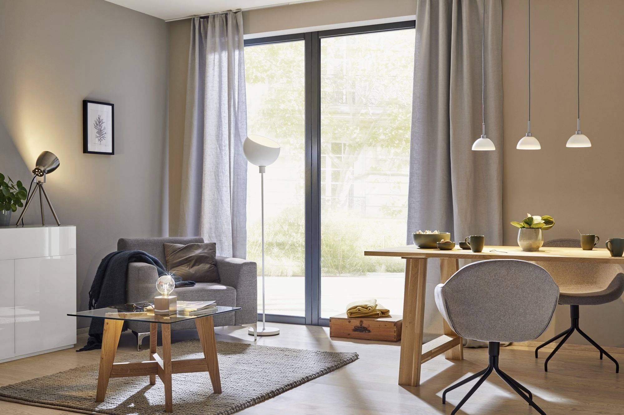 Moderná obývačka s sivou sedačkou, skleným stolíkom, jedálenským stolom a stoličkami. Veľké okná prepúšťajú veľa denného svetla.