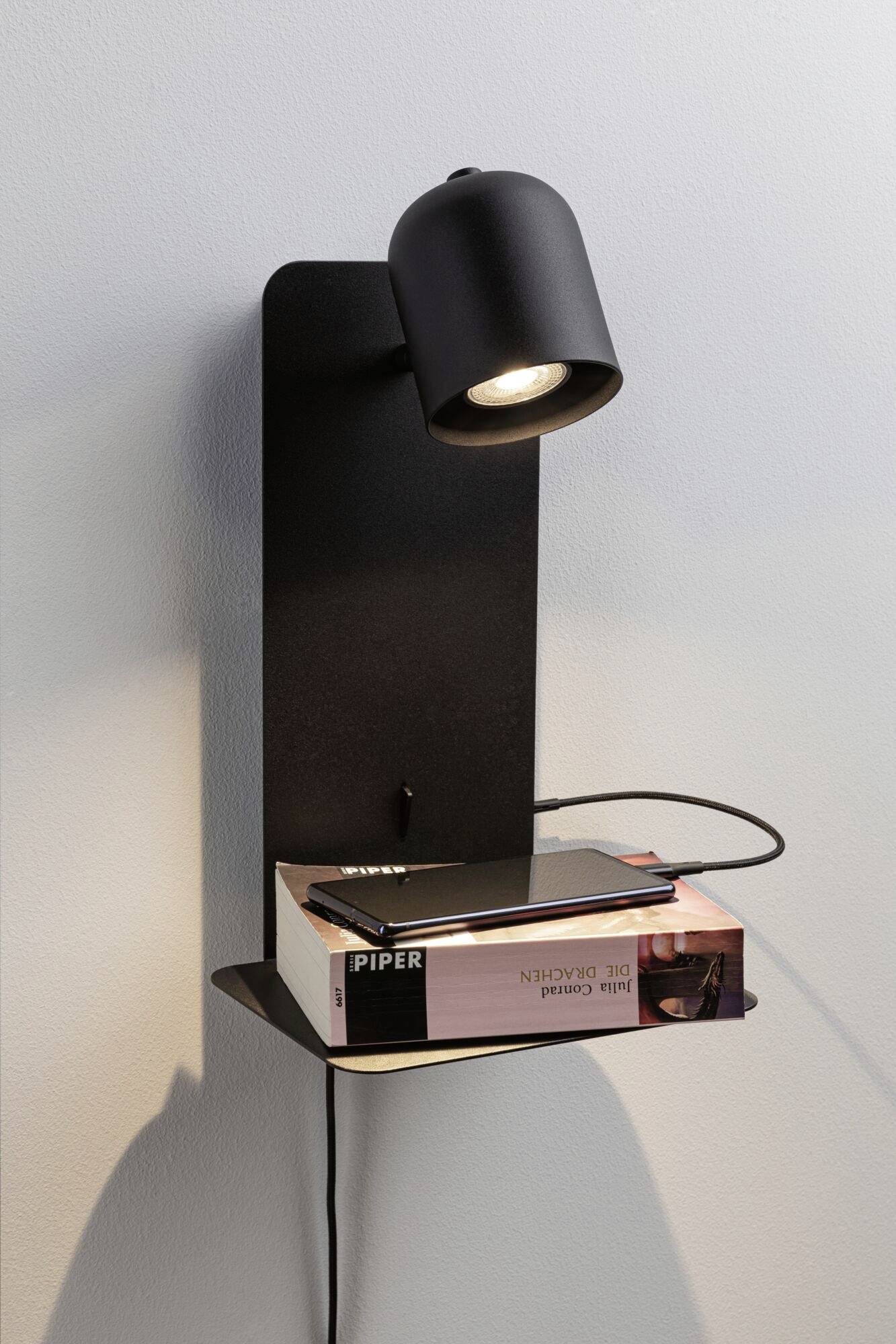 Čierna nástenná lampa so zabudovanou poličkou, na ktorej leží kniha a smartfón. Svetlo osvetľuje predmety.