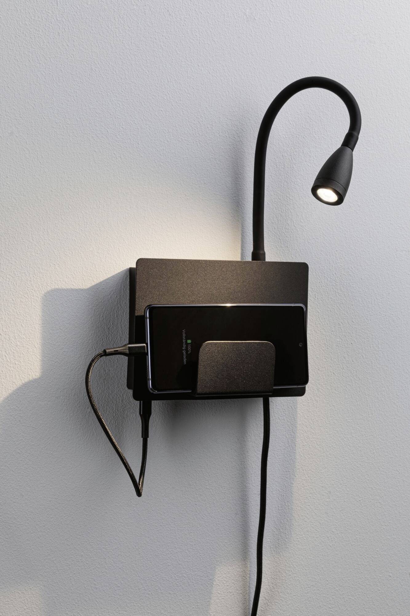 Mobilný telefón je pripevnený k nabíjačke namontovanej na stene. Nad ním sa nachádza flexibilná čítacia lampa.