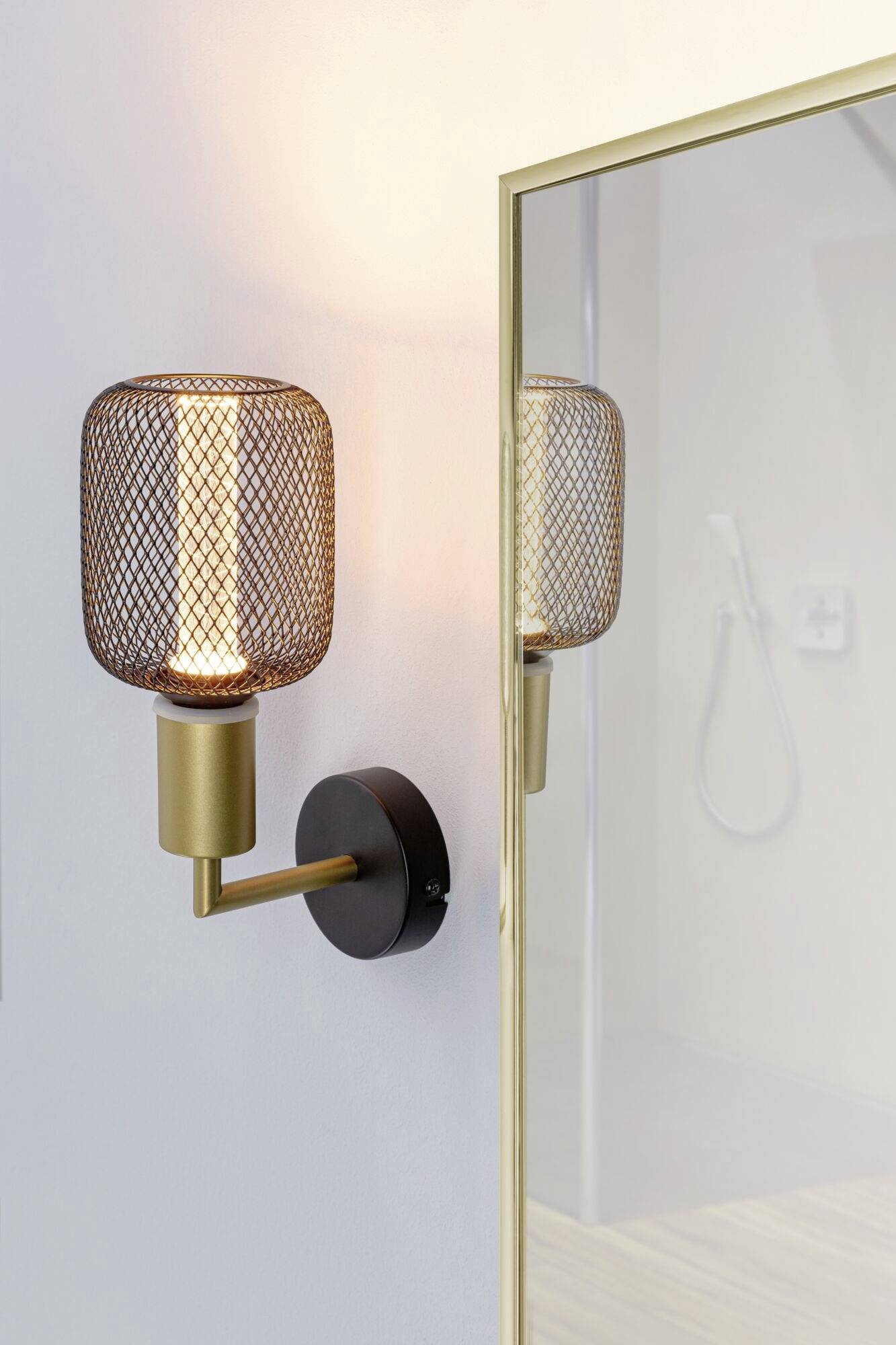 Zlatá nástenná lampa s mriežkovým dizajnom svieti vedľa zrkadla v modernej kúpeľni so sprchou v pozadí.