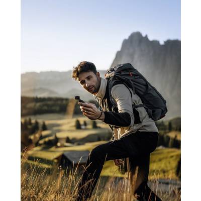 Muž s batohom drží fotoaparát v hornatej krajine. Ranné slnko osvecuje lúky a hory v pozadí.