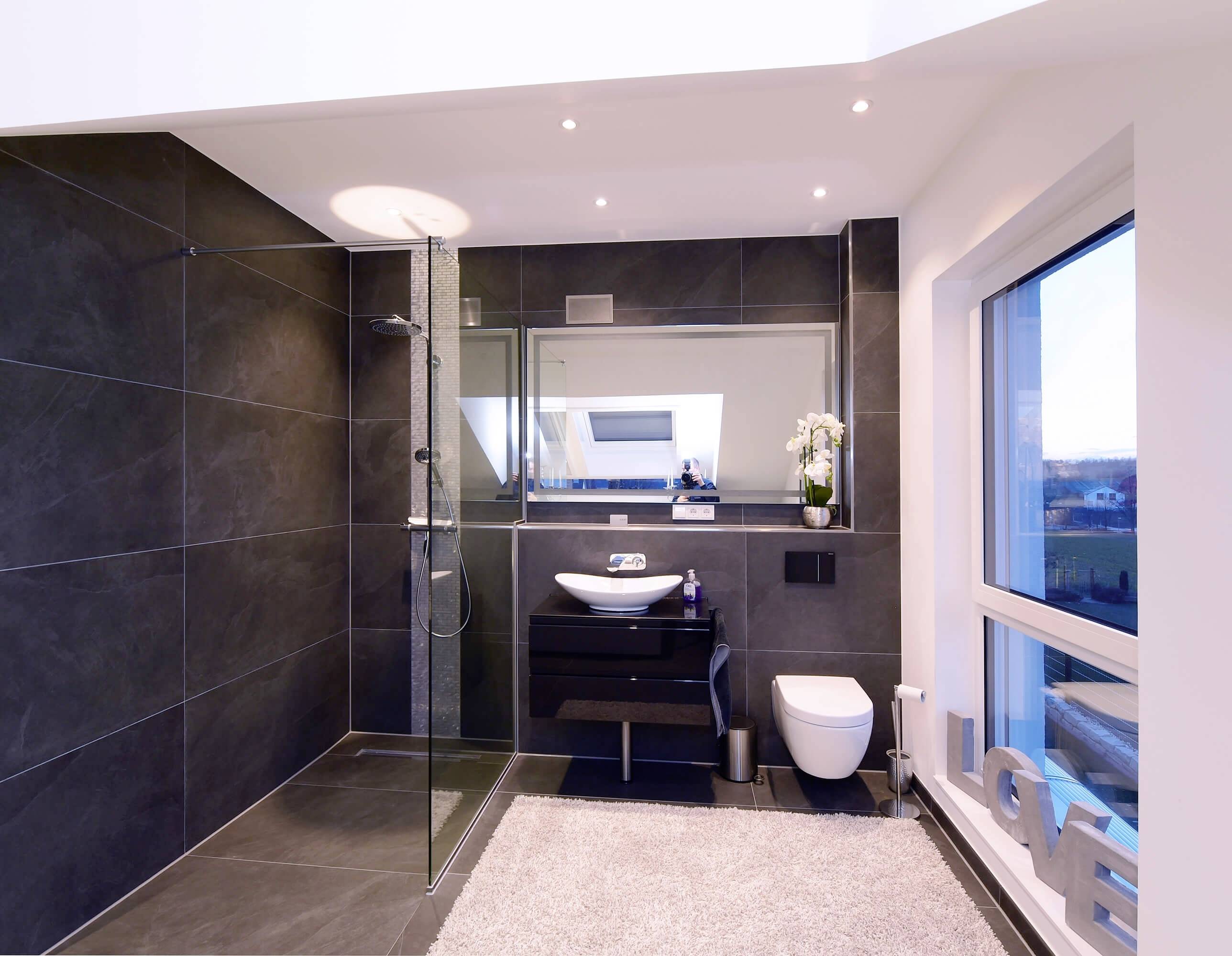 Moderná kúpeľňa so sprchou, umývadlom a toaletou. Tmavé obklady a veľké okno prepúšťajú množstvo denného svetla.