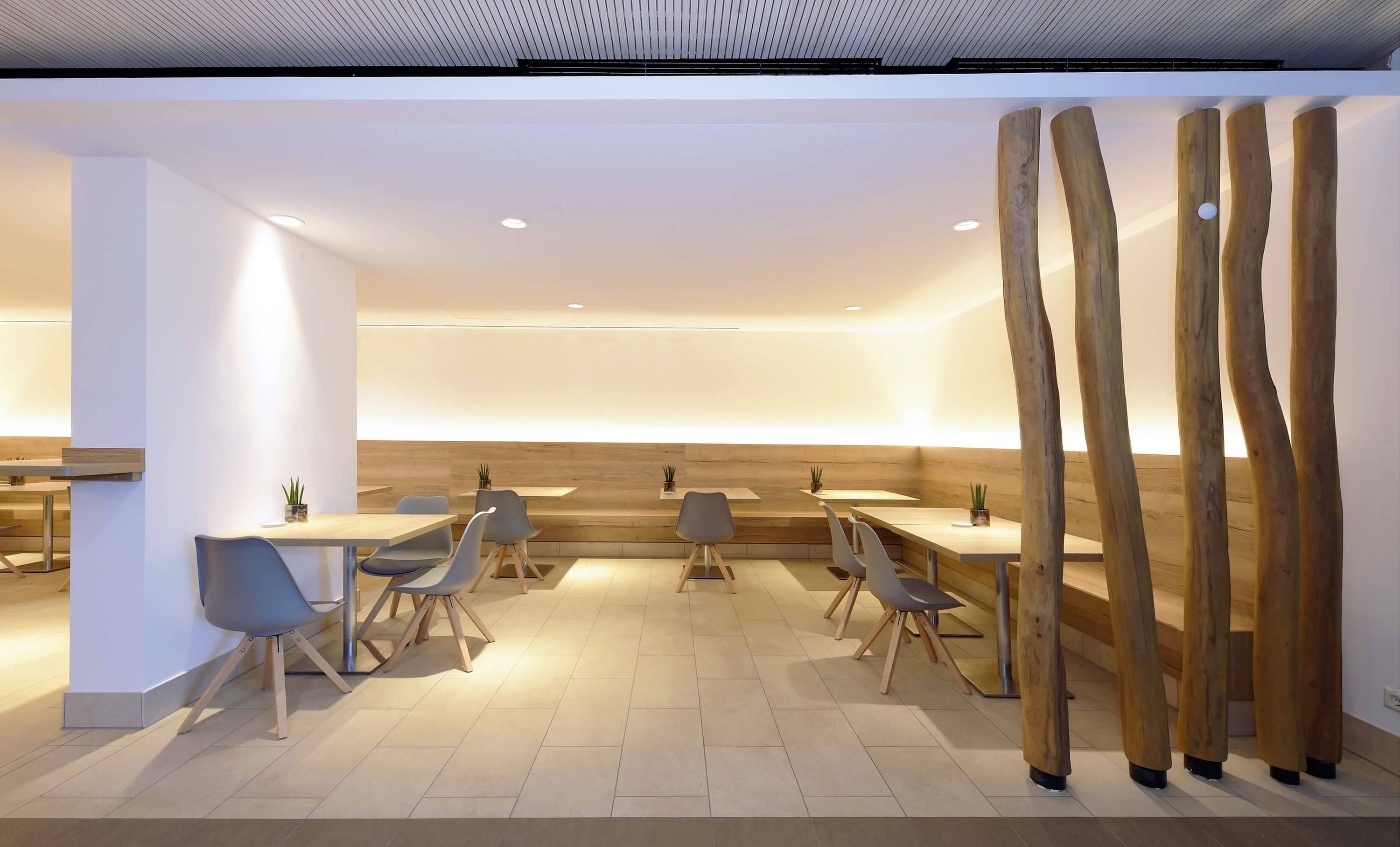 Moderne café so dreveným nábytkom a sivými stoličkami. Vľavo sú malé stolíky so stoličkami, vpravo väčšie lavice. Tlmené osvetlenie.