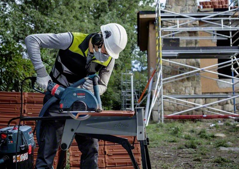 Robotník v ochrannej prilbe a bezpečnostnom oblečení používa elektrický nástroj na rezanie potrubia na stavenisku. V pozadí je vidieť lešenie pri budove.