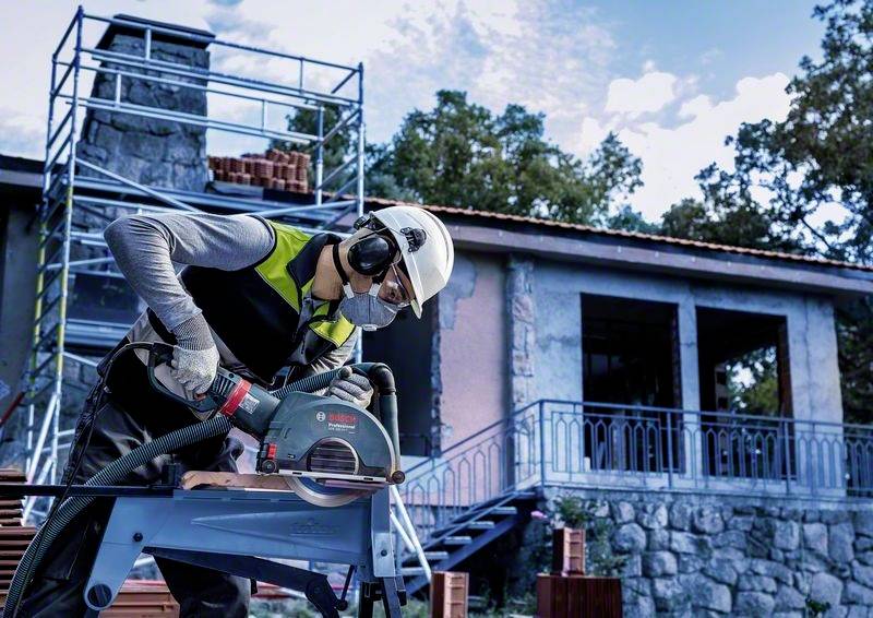Robotník v ochrannom oblečení obsluhuje elektrickú pílu pred budovou vo výstavbe so žeriavom v pozadí.