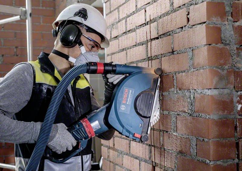 Robotník v ochranných okuliaroch, helme a maske upravuje tehlovú stenu pomocou elektrickej frézy.