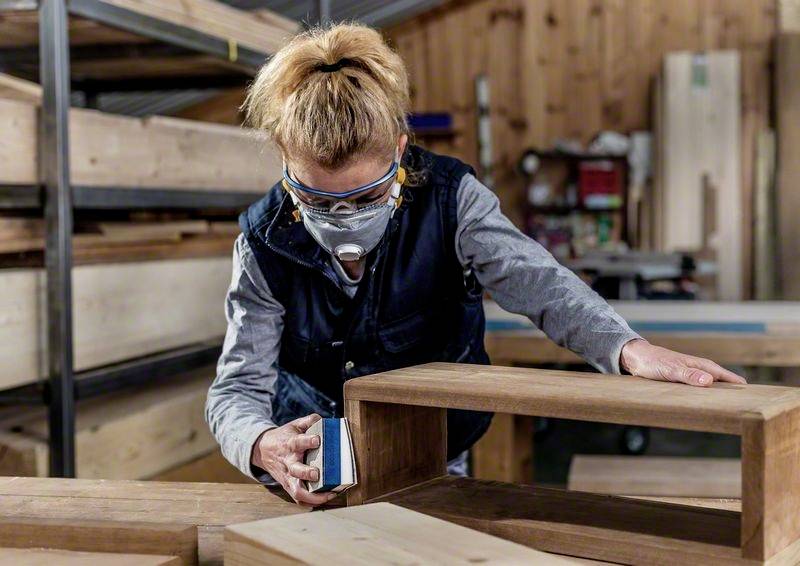 Osoba brúsi drevo v dielni. Má na sebe ochranné okuliare a respiračnú masku. V pozadí sú viditeľné drevené police.