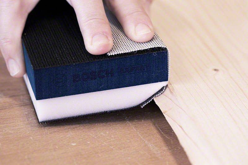 Ruka drží brúsnu špongiou na dreve, aby vyhladila povrch. Špongia zobrazuje logo známeho výrobcu náradia.