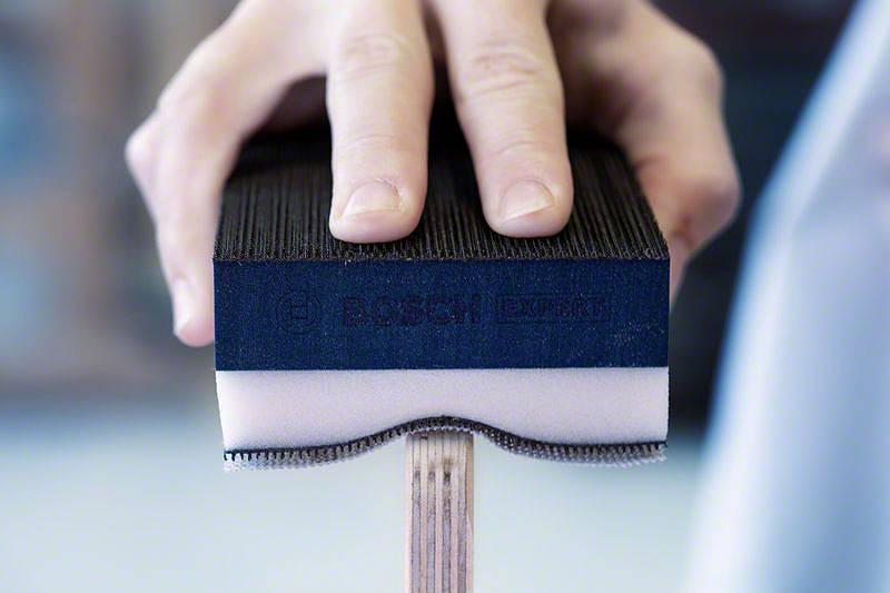 Jedna ruka drží brúsnu hubku Bosch, ktorá sa používa na drevenej doske. Hubka je dvojfarebná: čierna a biela.