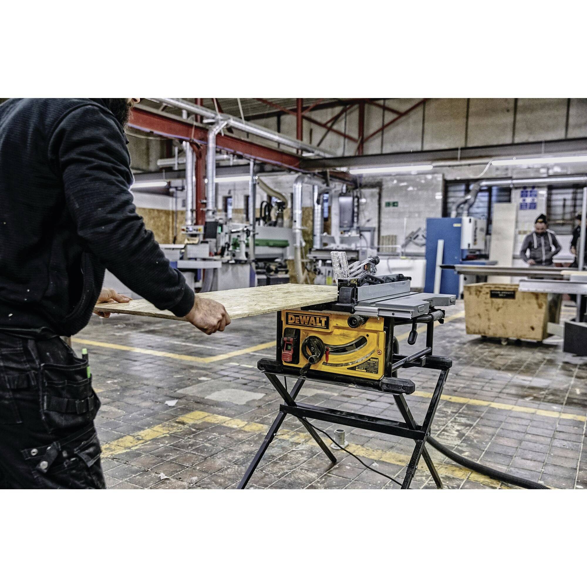 Osoba pracuje v dielni vedľa stolnej kotúčovej píly na stojane. V pozadí sú viditeľné stroje a drevené materiály.