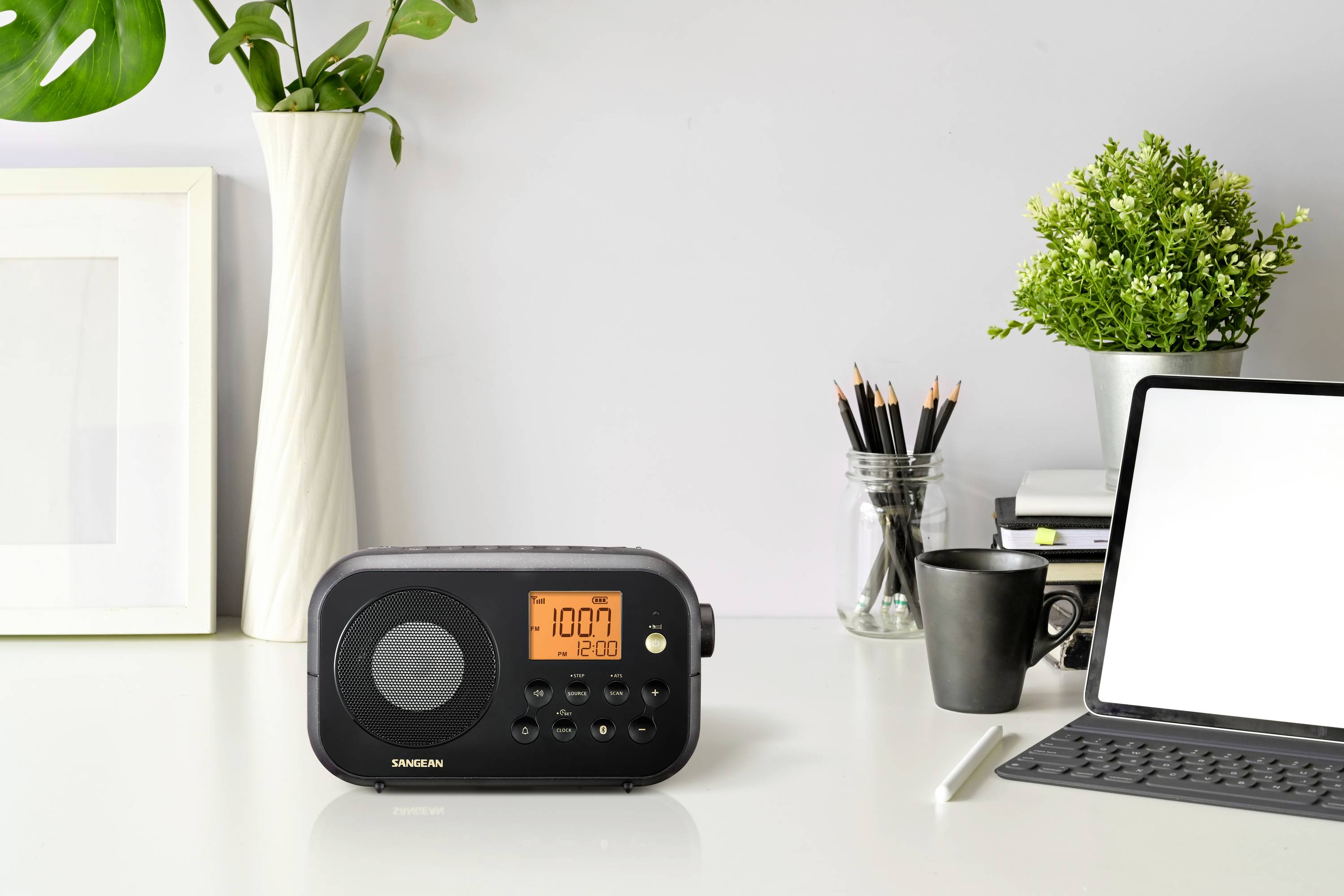 Rádio s osvetleným displejom ukazuje '10:07'. Stojí na stole vedľa laptopa, šálky a rastlín.
