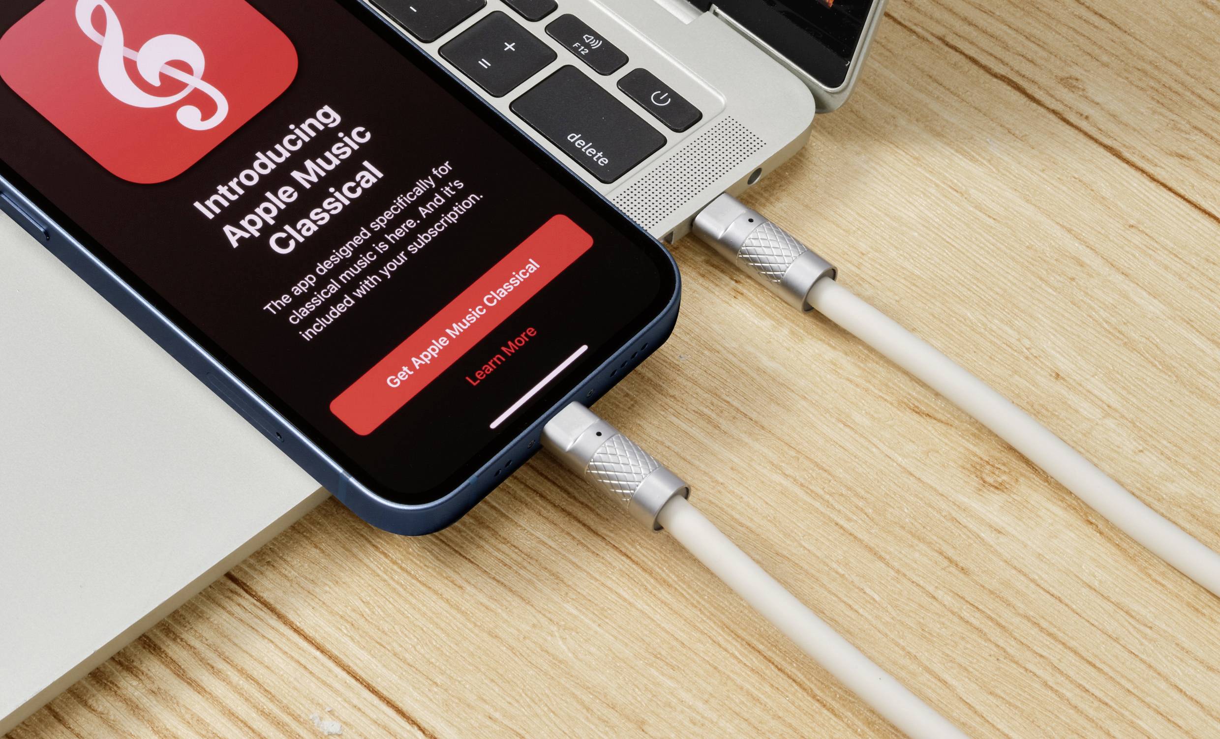 Smartfón zobrazuje oznámenie 'Apple Music Classical' na obrazovke, pripojený k laptoplu na drevenom stole.