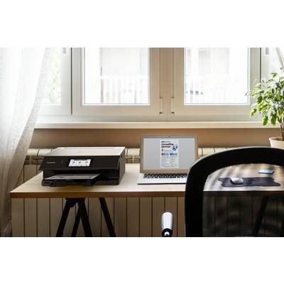 Pracovisko s laptopom, tlačiarňou a izbovou rastlinou na stole pred oknom. Laptop zobrazuje otvorenú webovú stránku.