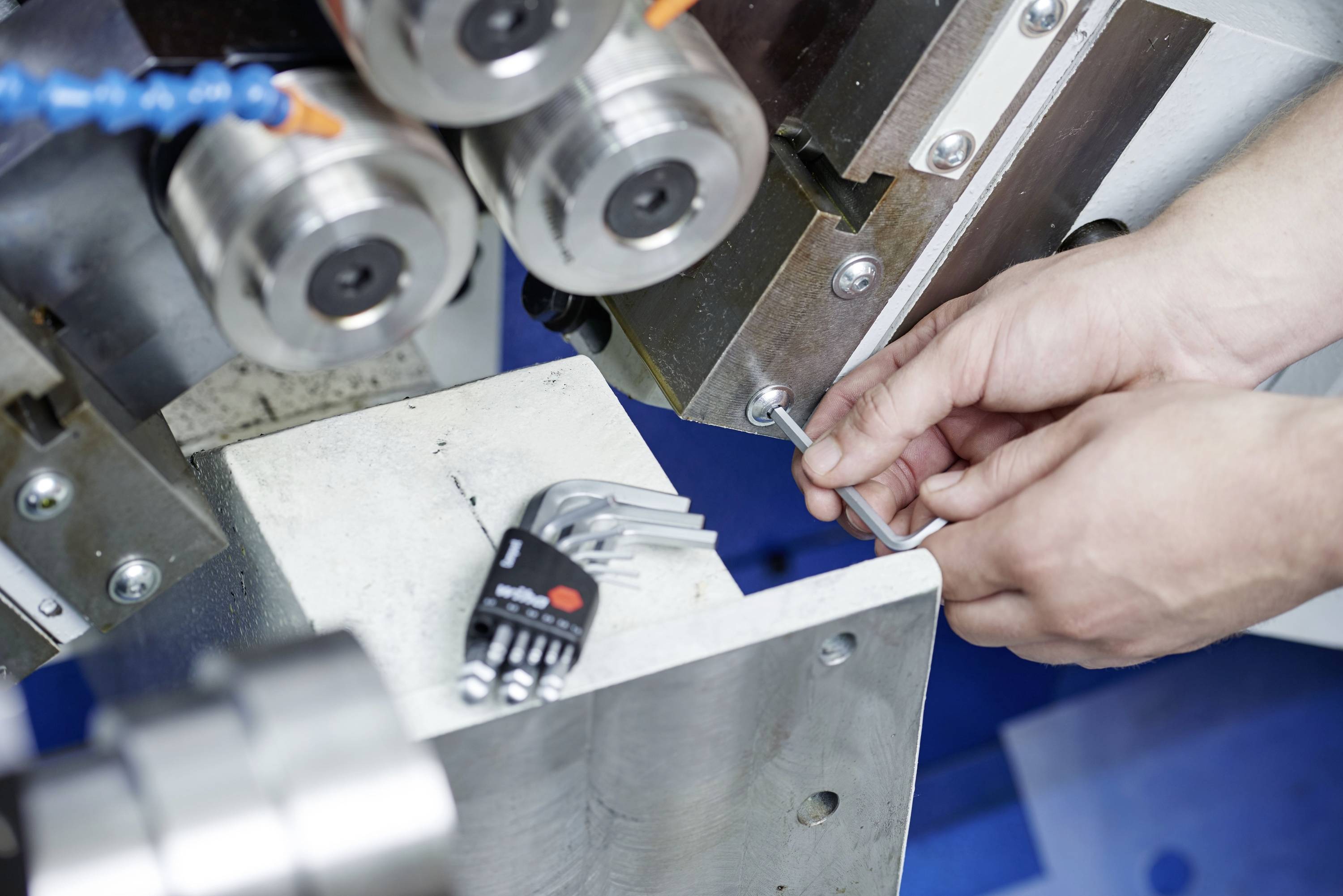 Niekoľko rúk nastavuje strojovú súčiastku pomocou imbusového kľúča. V popredí ležia ďalšie imbusové kľúče na stroji.