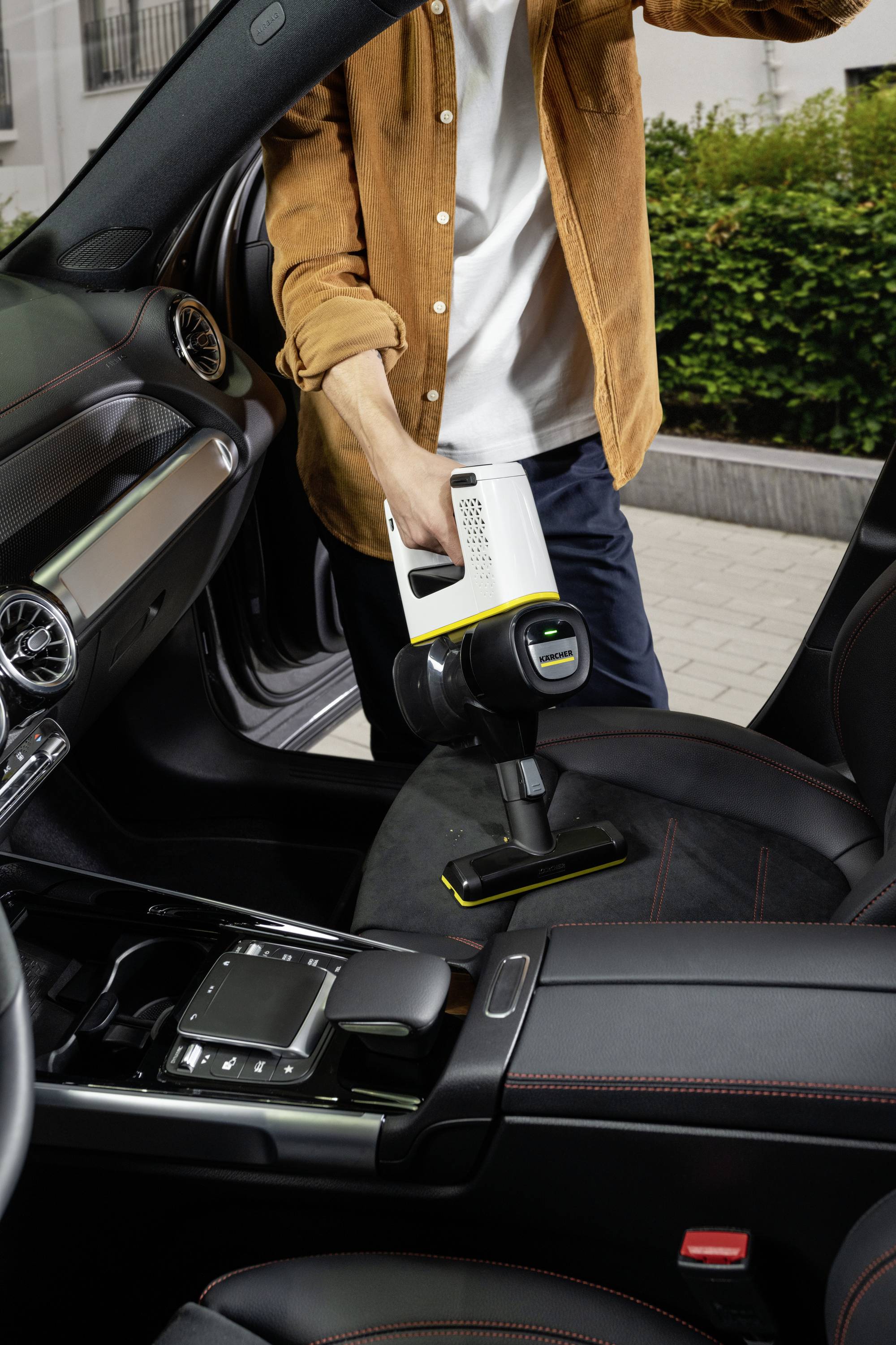 Osoba vysáva autosedadlo ručným vysávačom. Interiér auta je moderný s koženými detailmi.