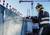 Robotník vŕta do betónovej steny na stavenisku. Má na sebe ochranný odev a slúchadlá. V pozadí je vidieť žeriav.