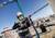 Stavebný robotník v ochrannej prilbe a respirátore vŕta výkonným príklepovým vŕtacím kladivom na stavenisku, obklopený žeriavom a štruktúrou budovy.