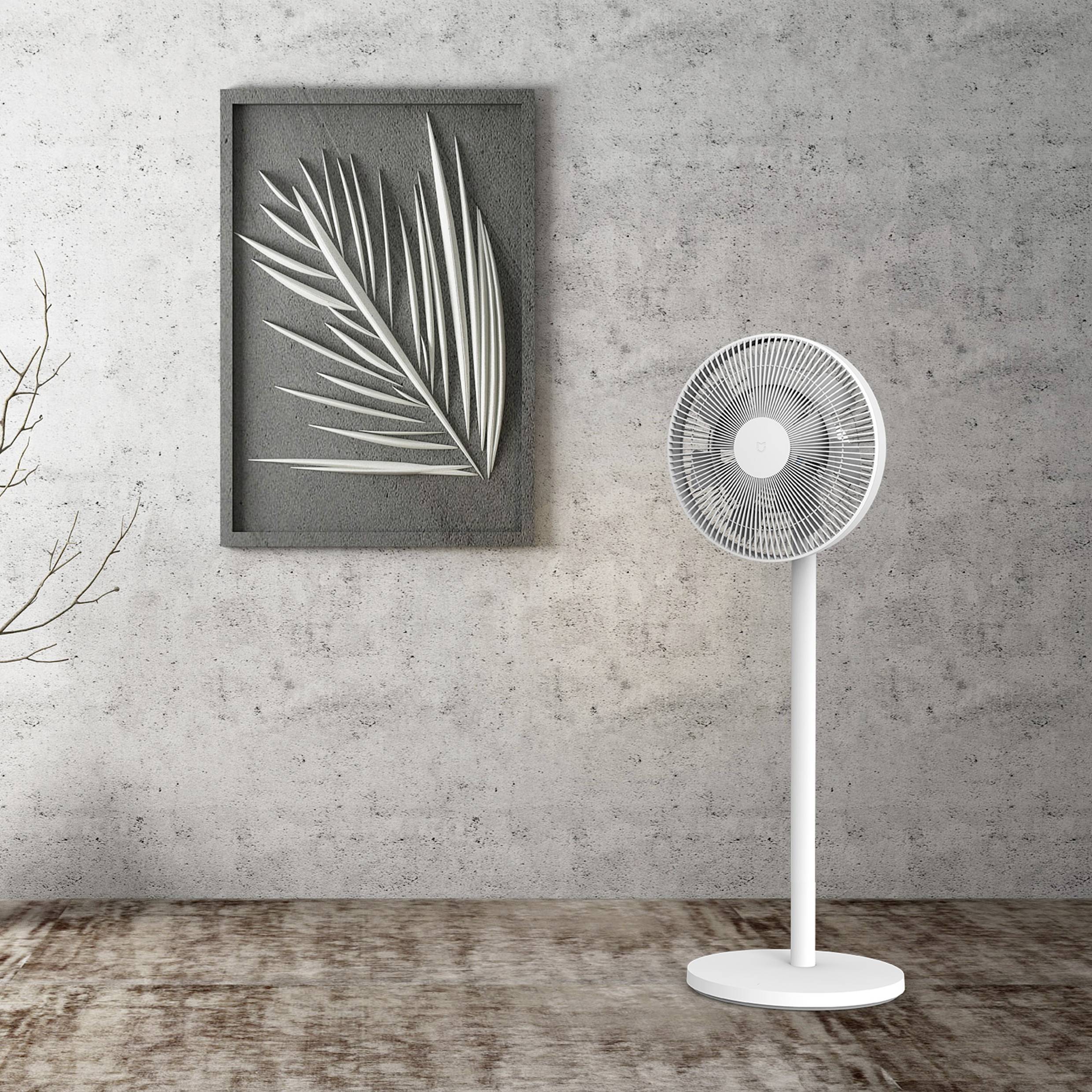 Biely stojanový ventilátor stojí v miestnosti s umeleckým dielom, ktoré zobrazuje štylizovanú reprezentáciu listov.