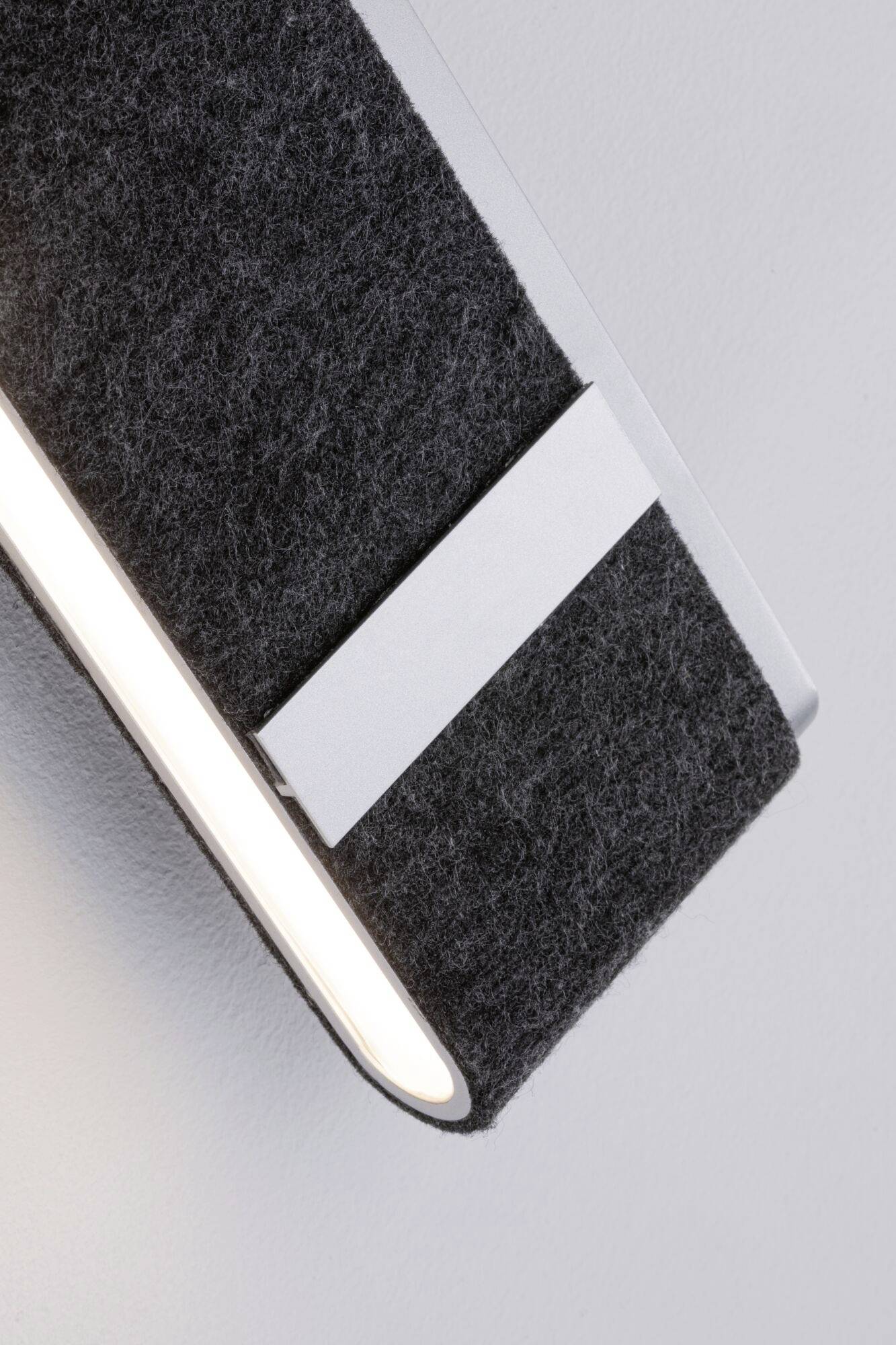 Moderná nástenná lampa s šedým plsteným krytom a úzkym svetelným pruhom, namontovaná na svetlej stene.