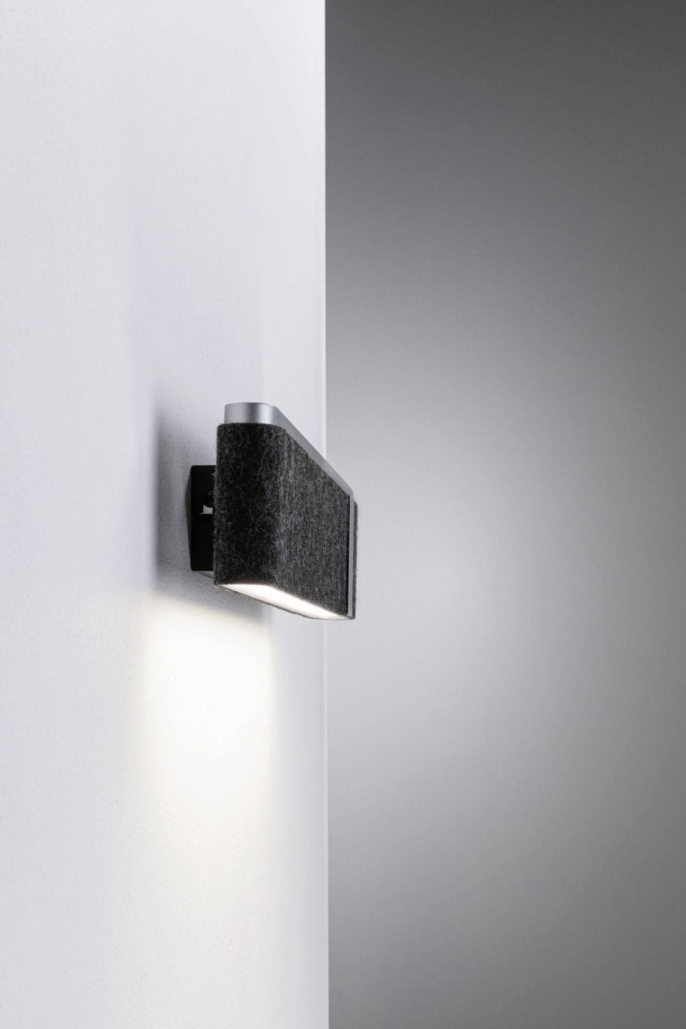 Kovená nástenná lampa s čiernym textilným tienidlom, ktorá jemne osvecuje bielu stenu. Moderný, minimalistický dizajn.