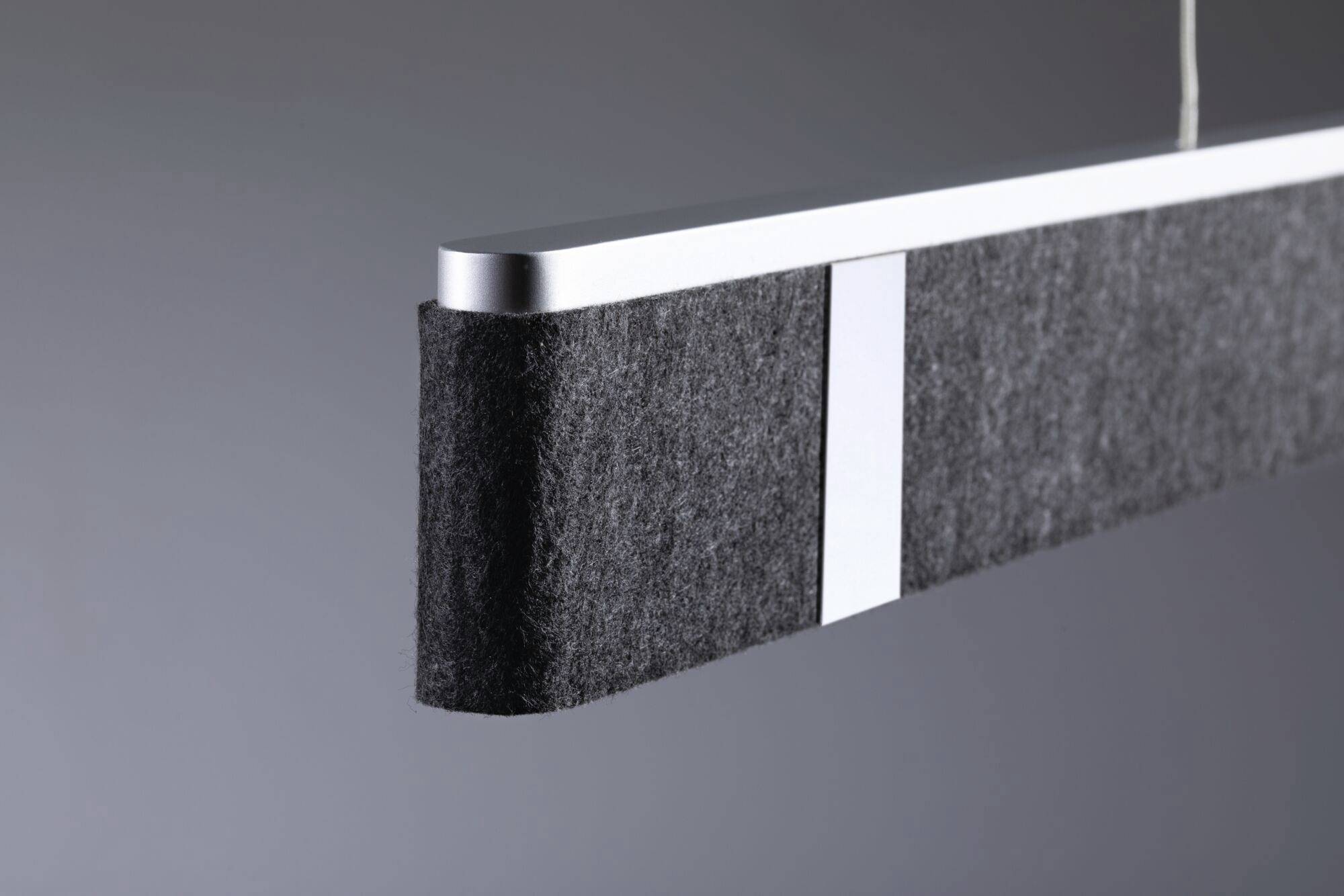 Sivý, obdĺžnikový závesný luster s textilnými prvkami na oboch koncoch, pred neutrálnym pozadím.