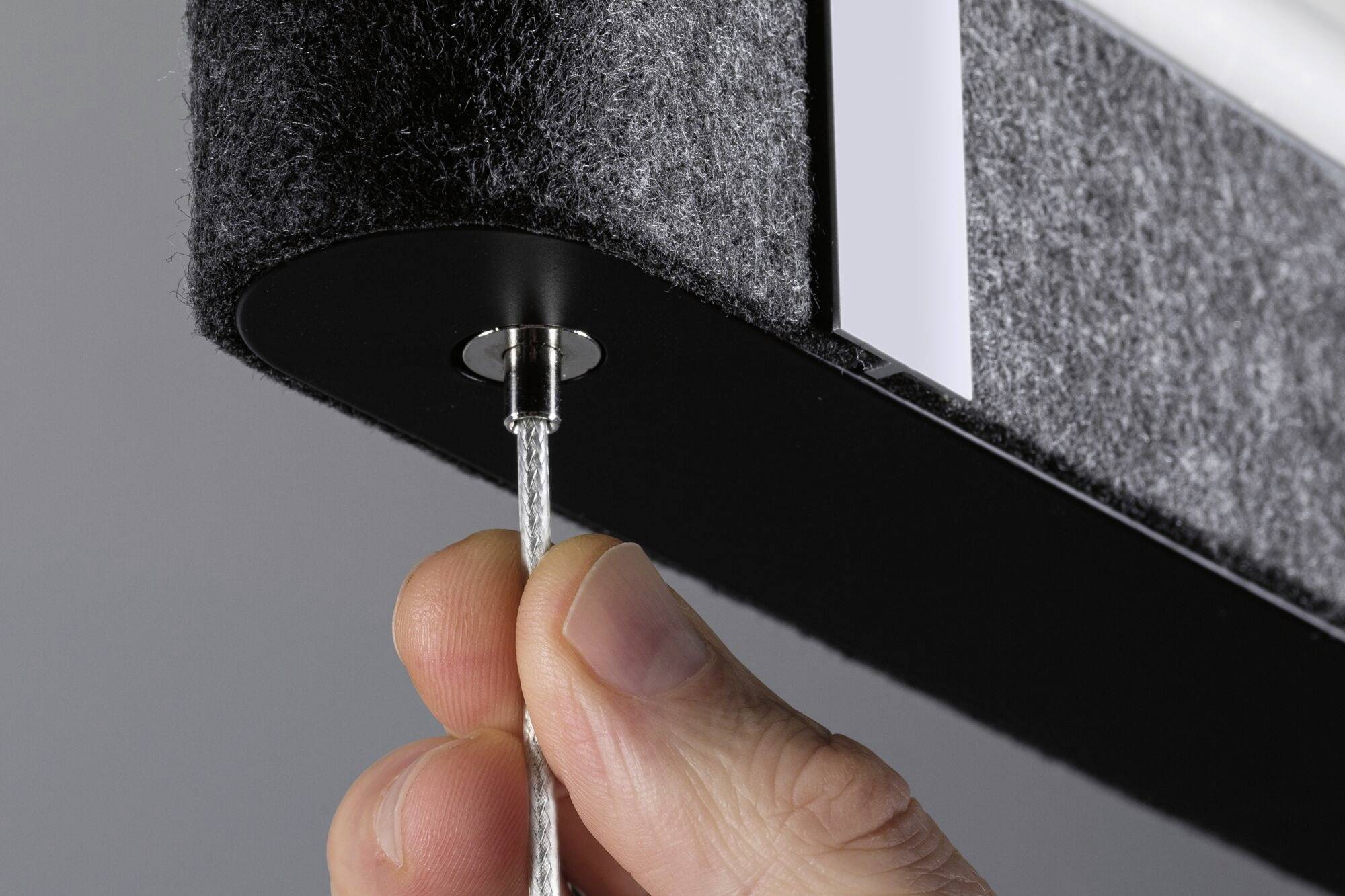 Jedna ruka nastavuje oceľové lanko na čiernej, plsteným poťahom pokrytej stropnej lampe, ktorá je namontovaná na strope.