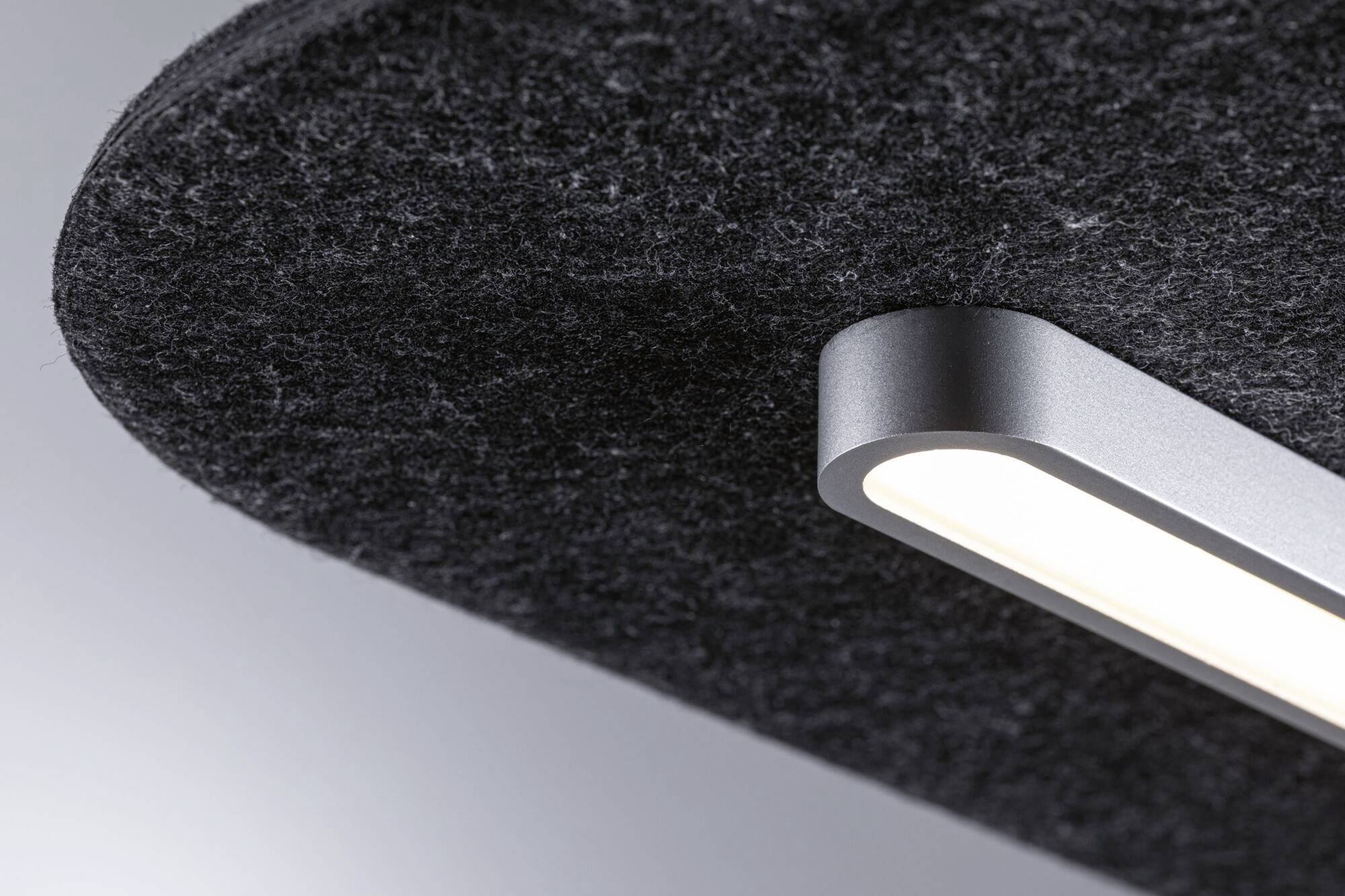 Moderná, podlhovastá stropná lampa s čiernym krytom osvetľuje šedý štruktúrovaný povrch zavesený na strope.