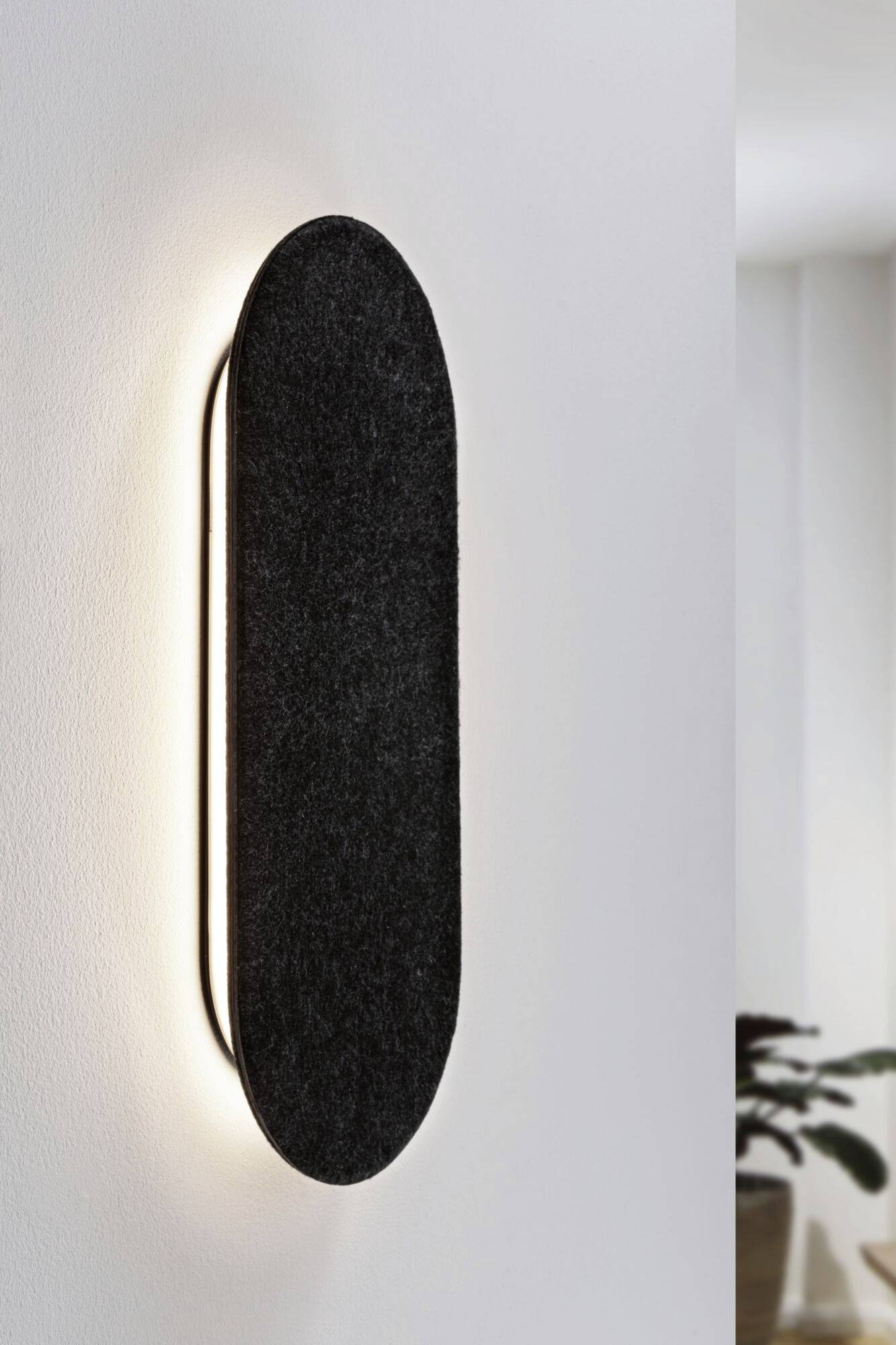 Nástenná lampa v modernom dizajne s čiernym, oválnym textilným tienidlom, jemne osvecuje bielu stenu v minimalistickom priestore.