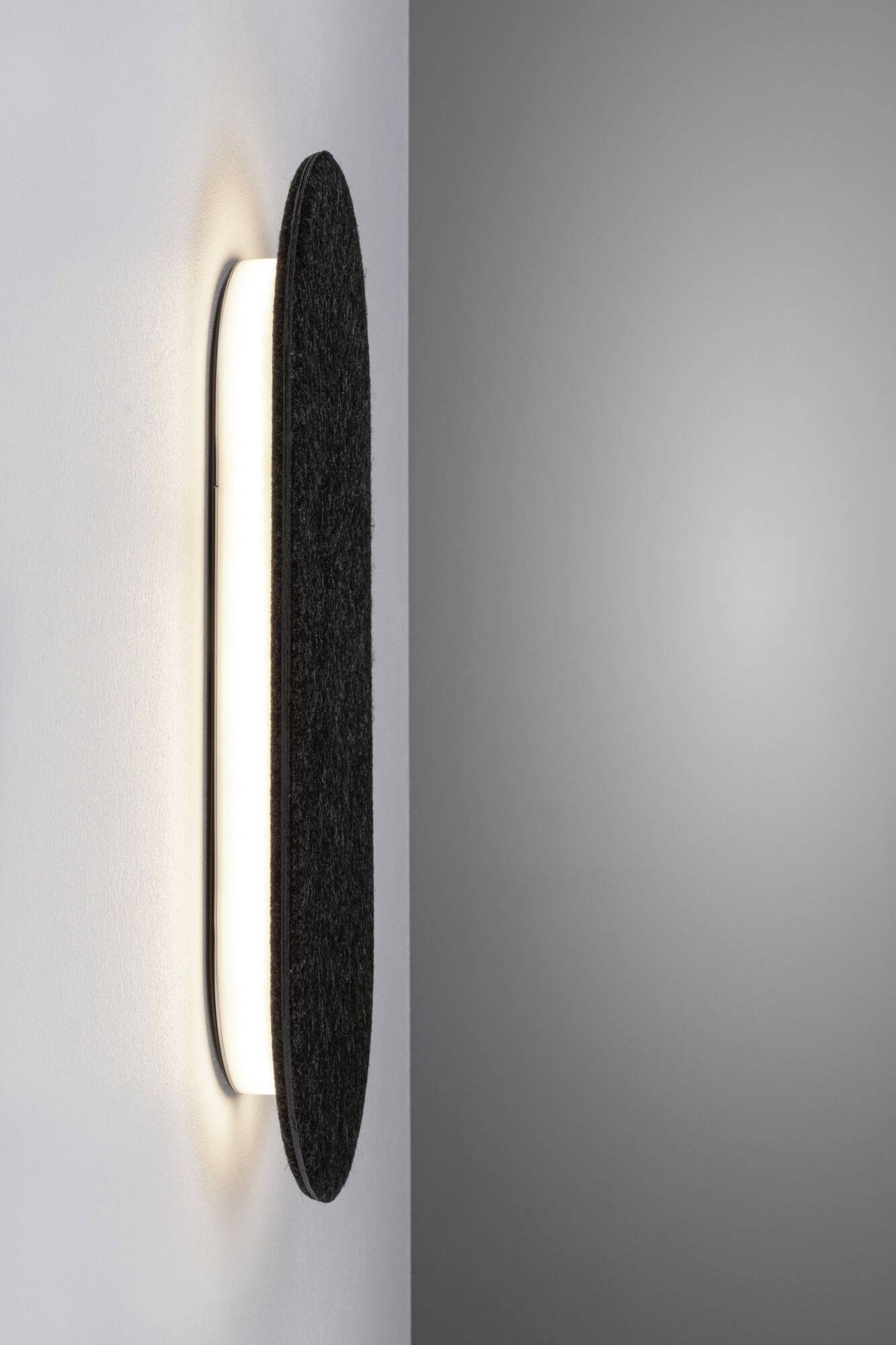 Nástenná lampa s oválnym čiernym tienidlom na sivej stene, podsvietená, vytvára moderný priestor.