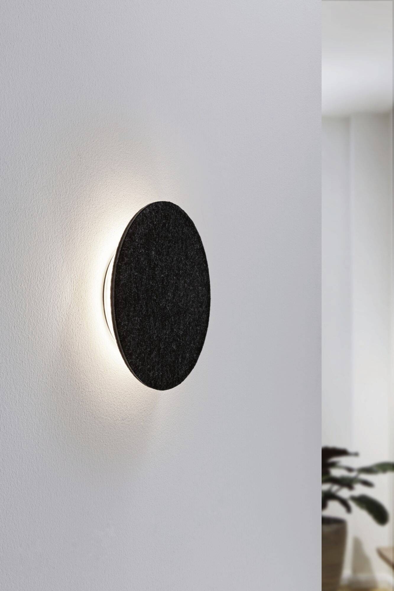 Čierne, okrúhle nástenné svietidlo s nepriamym osvetlením, namontované na bielej stene. V pozadí rozmazaný, moderný interiér.