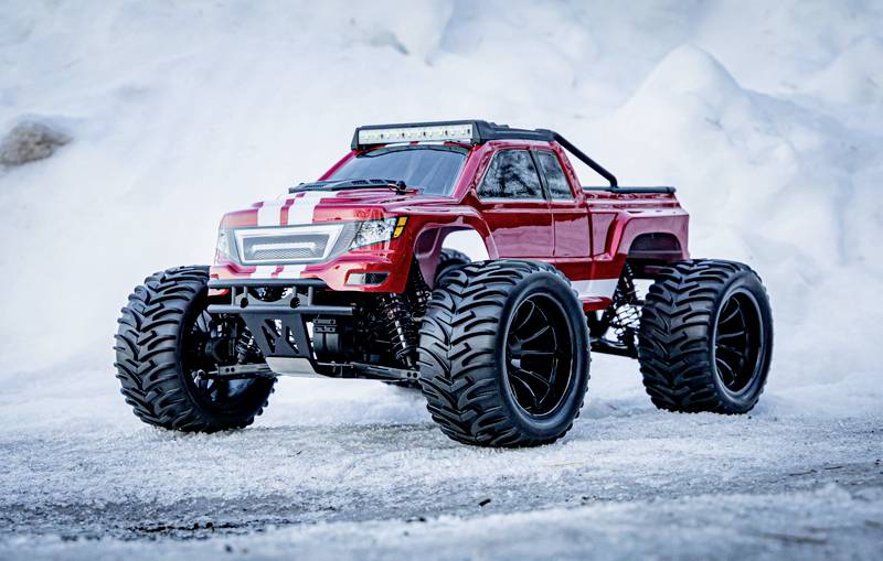 Červený model monster trucku na diaľkové ovládanie stojí na zasneženom povrchu. Veľké pneumatiky, sneh v pozadí.
