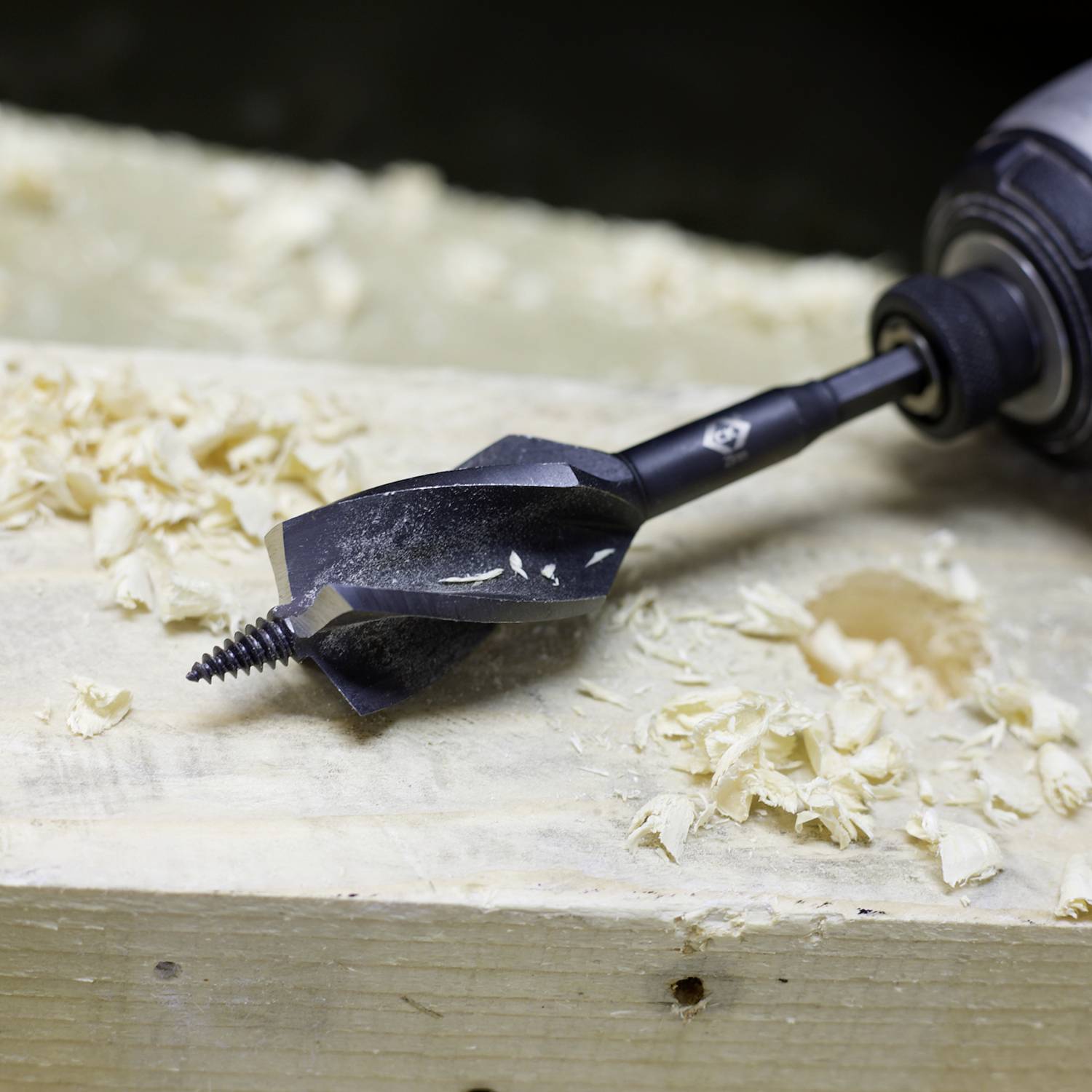 Vŕtačka s nástavcom na zemné vŕtanie na drevenej doske, obklopená trieskami dreva.