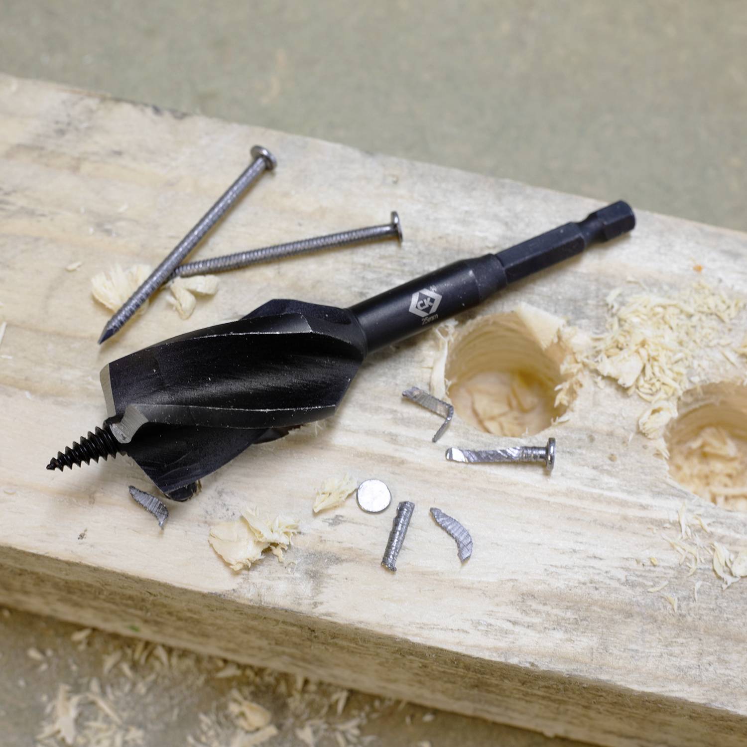 Vrták vedľa skrutiek na drevenej doske s dvomi vyvŕtanými otvormi, obklopený trieskami dreva.