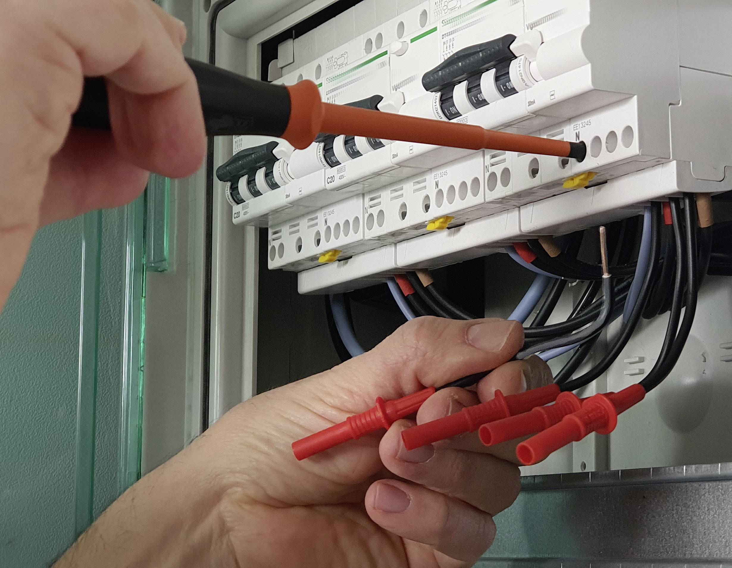 Osoba opravuje alebo inštaluje poistkovú skrinku, drží červené káble a používa skrutkovač na prácu s elektrickými obvodmi.