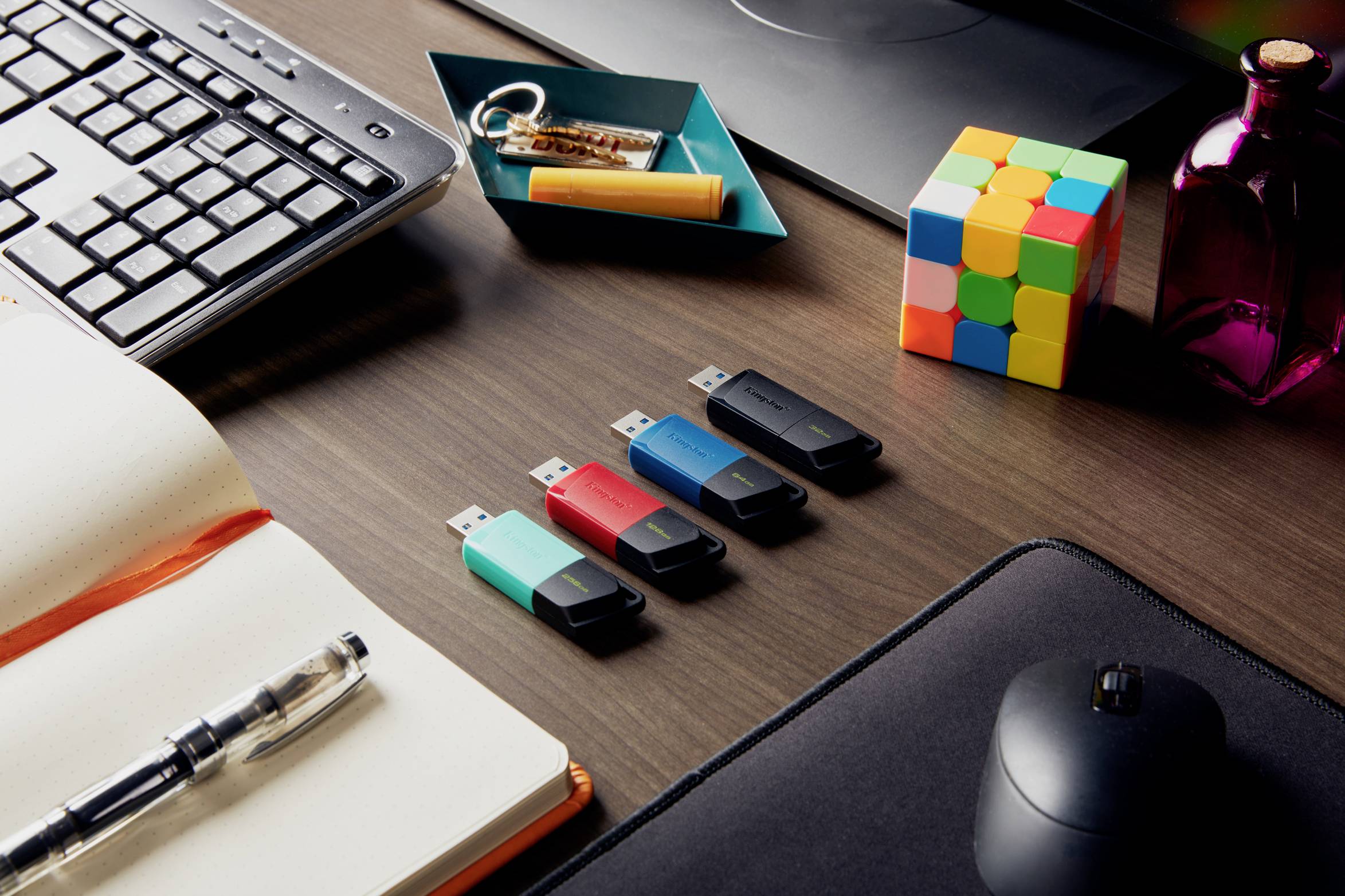USB kľúče v rôznych farbách ležia na písacom stole, obklopené klávesnicou, myšou, poznámkovým blokom, perom a kockou.