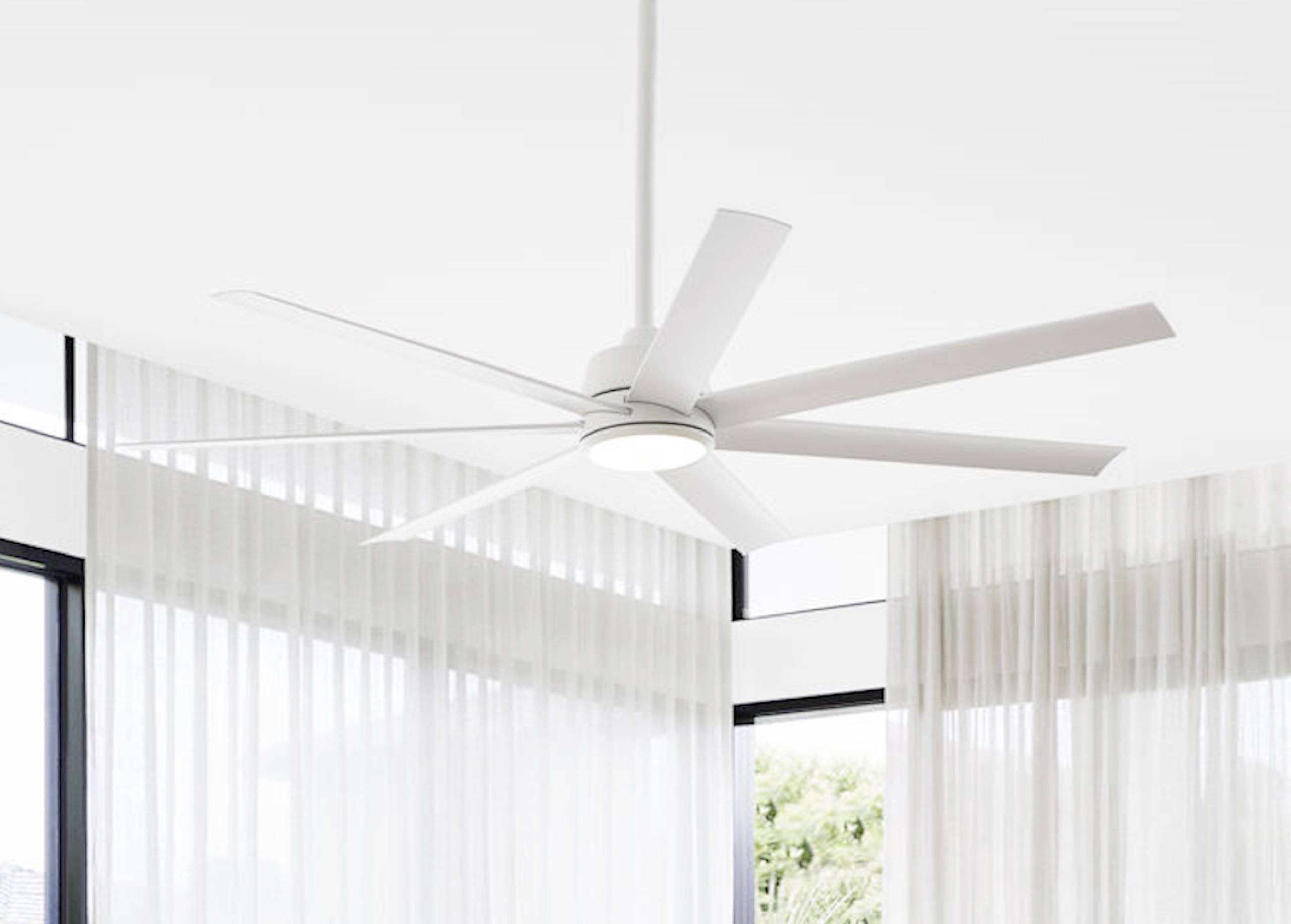 Biely stropný ventilátor s viacerými krídlami je nainštalovaný na strope svetlej miestnosti s veľkými oknami a polopriehľadnými záclonami.