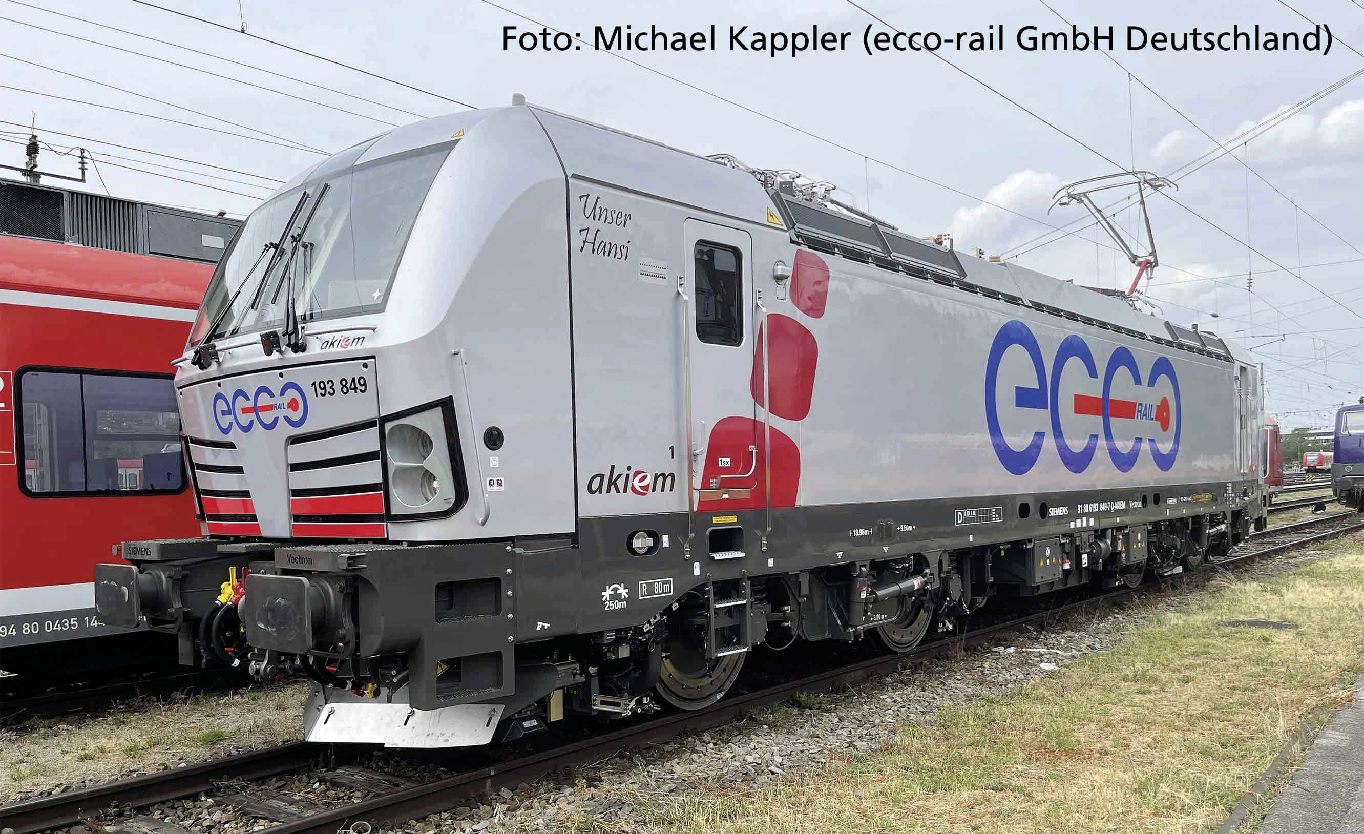 Sivá lokomotíva s červeným a modrým logom 'ecco'. Postranný nápis 'Náš Hans' a 'akiem'. Pohybuje sa po železničných koľajach pod elektrickým vedením.