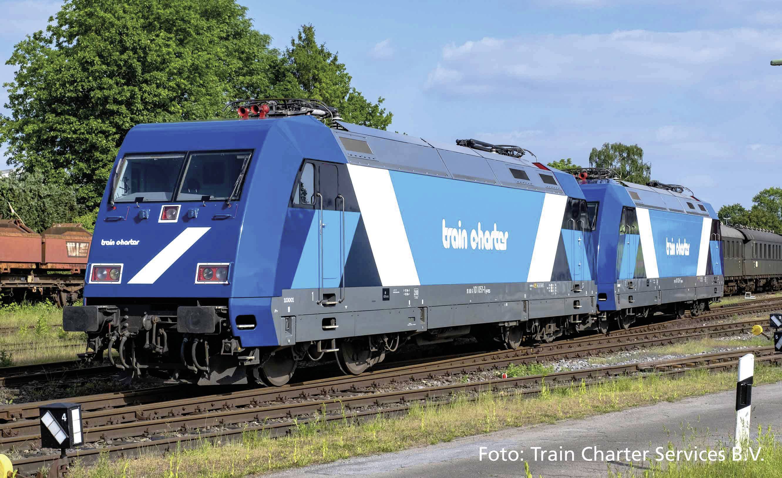 Dva modré vlaky s nápisom 'train charter' jazdia po koľaji vonku. Stromy a obloha sú v pozadí.
