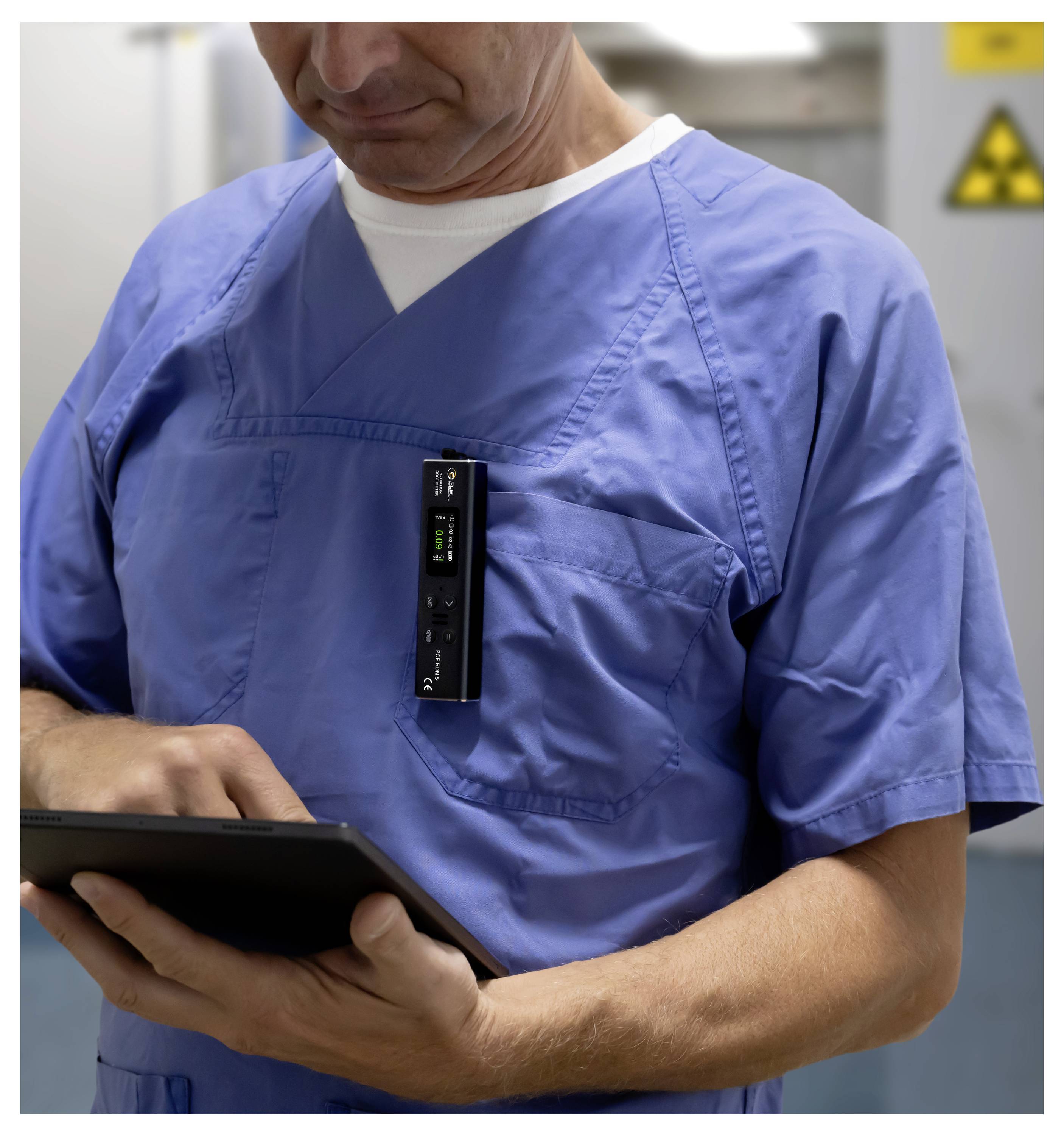 Zdravotnícky pracovník v modrých pracovných odevoch používa tablet s klipom pripevneným elektronickým zariadením na hrudi v nemocničnom prostredí.