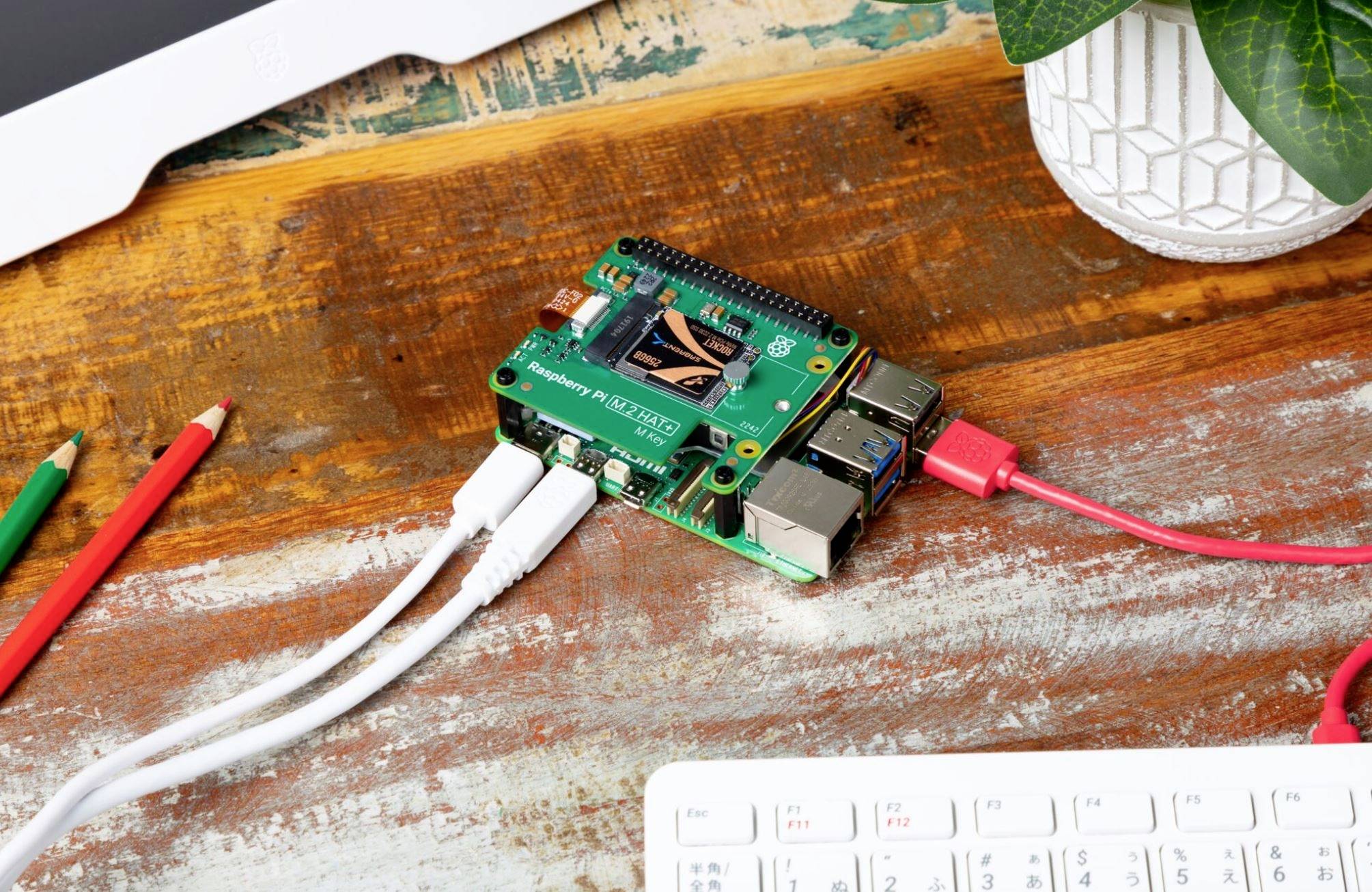 Na stole leží Raspberry Pi s pripojenými káblami vedľa ceruziek a rastliny, obklopený počítačovým vybavením.