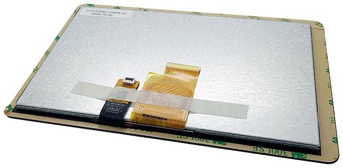 Plochý elektronický komponent s oranžovými konektormi, ležiaci na kartóne. Pravdepodobne obrazovka alebo modul.