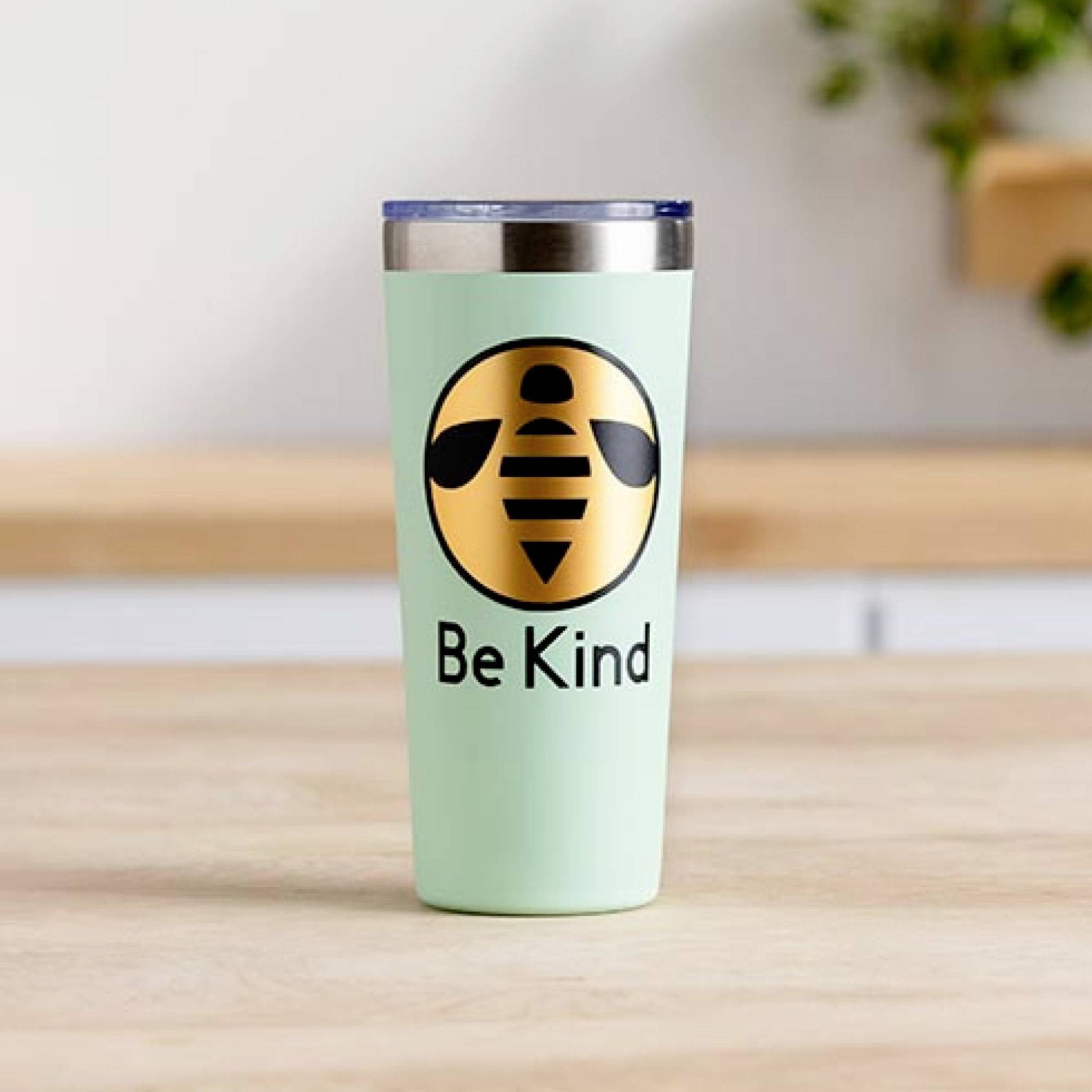 Mätový pohár so štylizovaným logom včely a textom 'Be Kind' stojí na stole. V pozadí sú rozmazané kuchynské prvky.