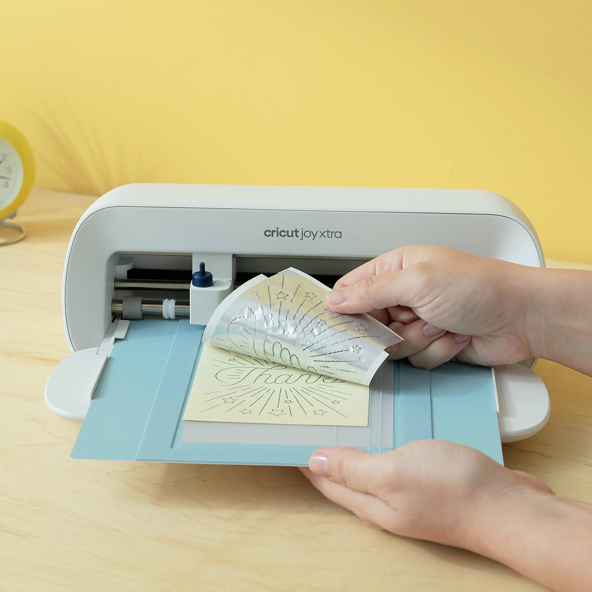 Ruka vyťahuje potlačenú fóliu z bieleho rezacieho zariadenia s nápisom 'cricut joy xtra'. Pozadie je v odtieňoch žltej.