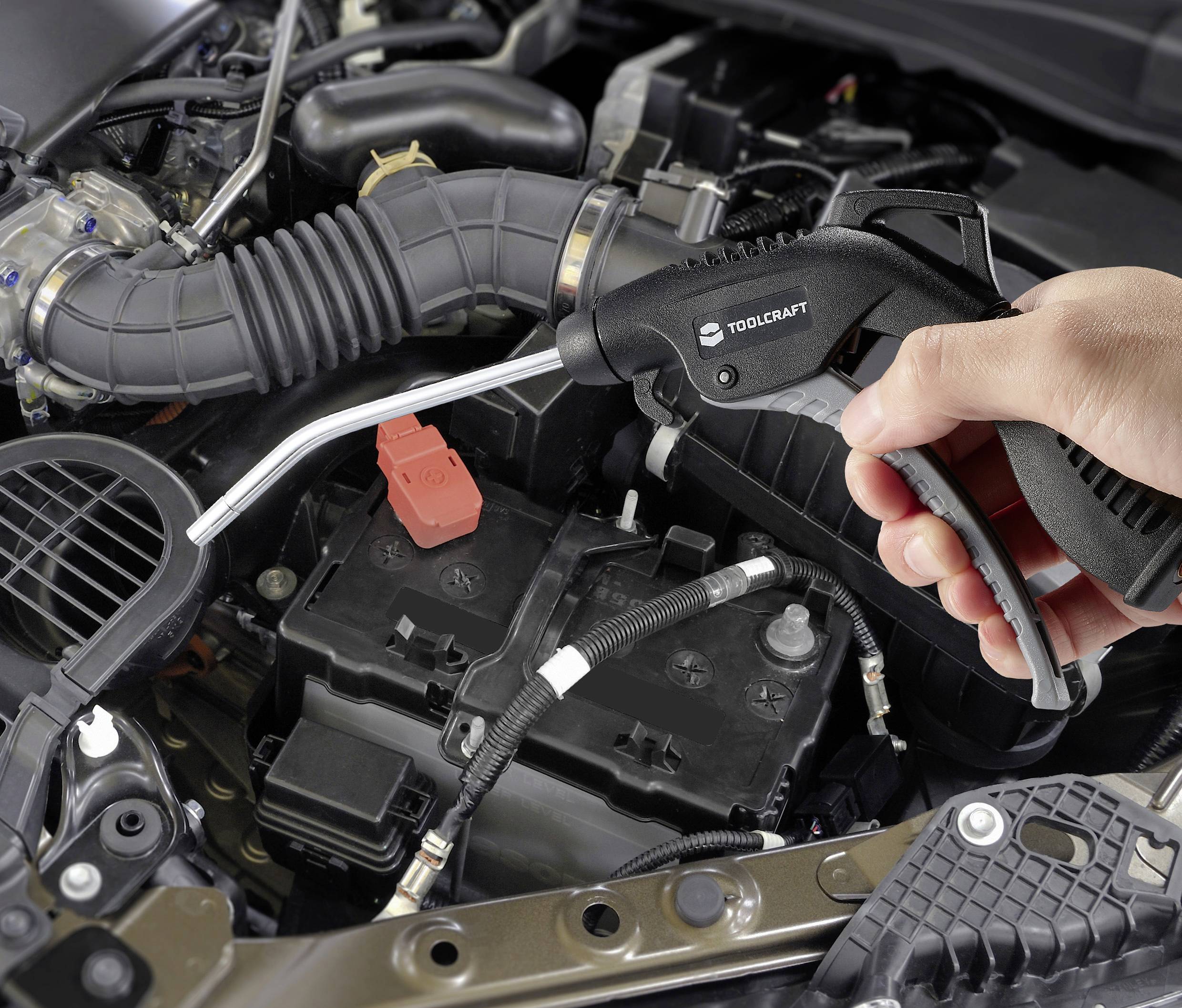 Jedna ruka drží pištoľ so stlačeným vzduchom nad otvorenou kapotou automobilu. Priestor motora je viditeľný v podrobných detailoch.