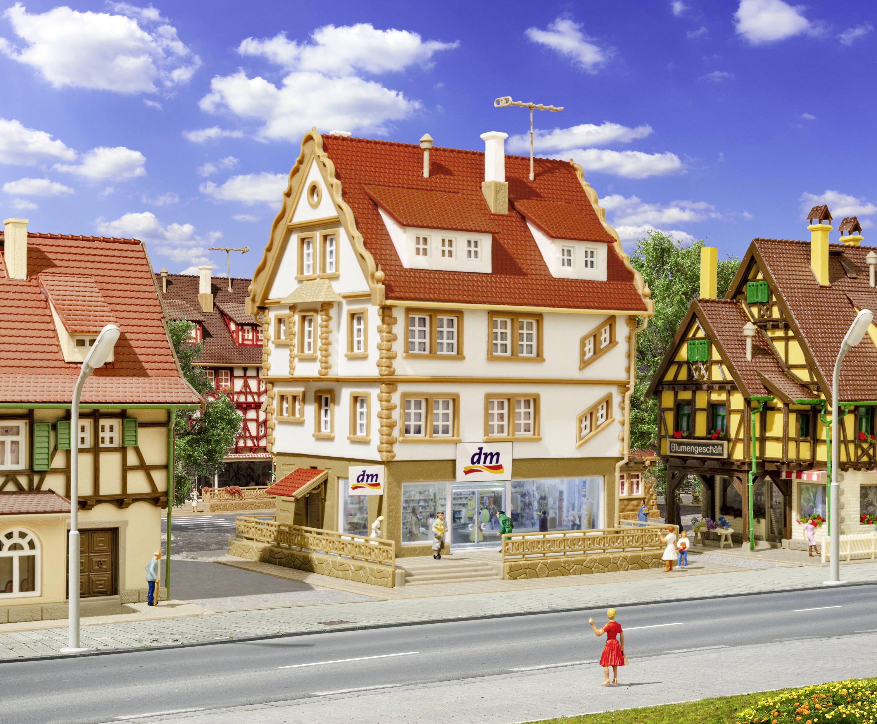 Miniatúrne mesto s hrázdenými domami, úzka ulička a ľudia. V popredí budova s logom 'dm'.