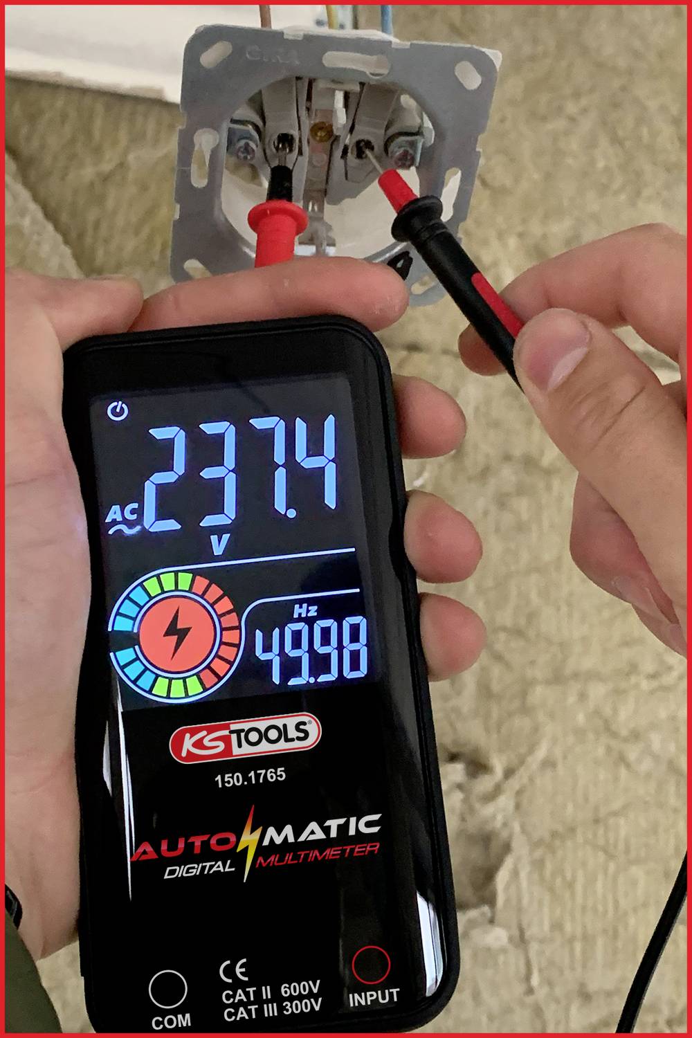 'Jedna ruka drží digitálny multimeter, ktorý zobrazuje 237,4 V striedavého napätia, pričom je pripojený do elektrickej zásuvky.'