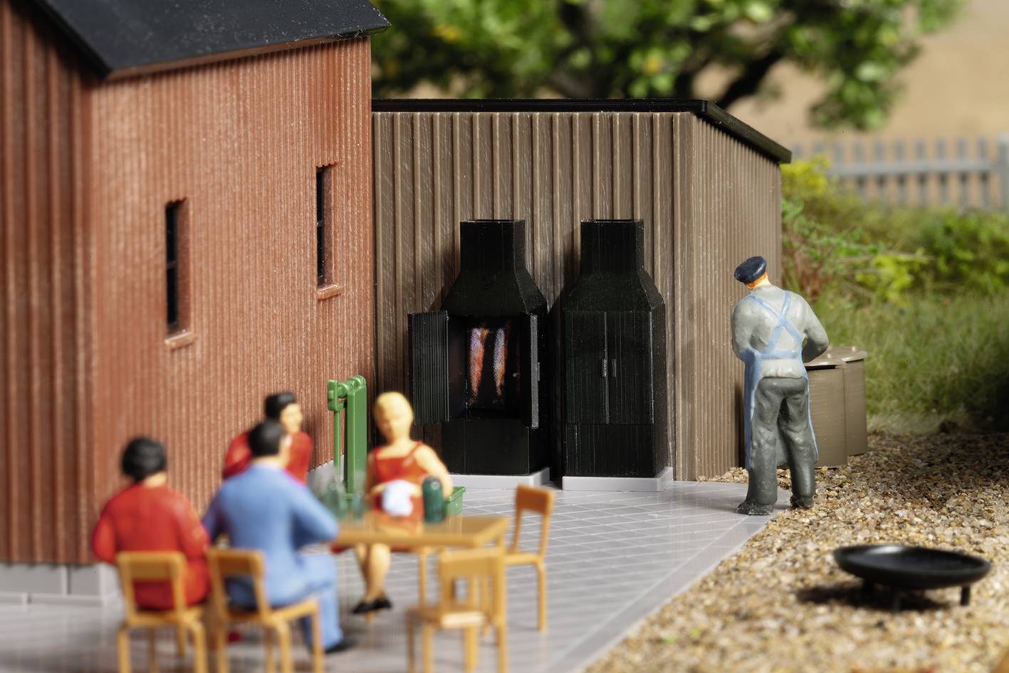 Modelová železnica - dioráma: Päť miniatúrnych figúrok vonku; traja sedia pri stole, jeden stojí pred grilom, drevené budovy v pozadí.